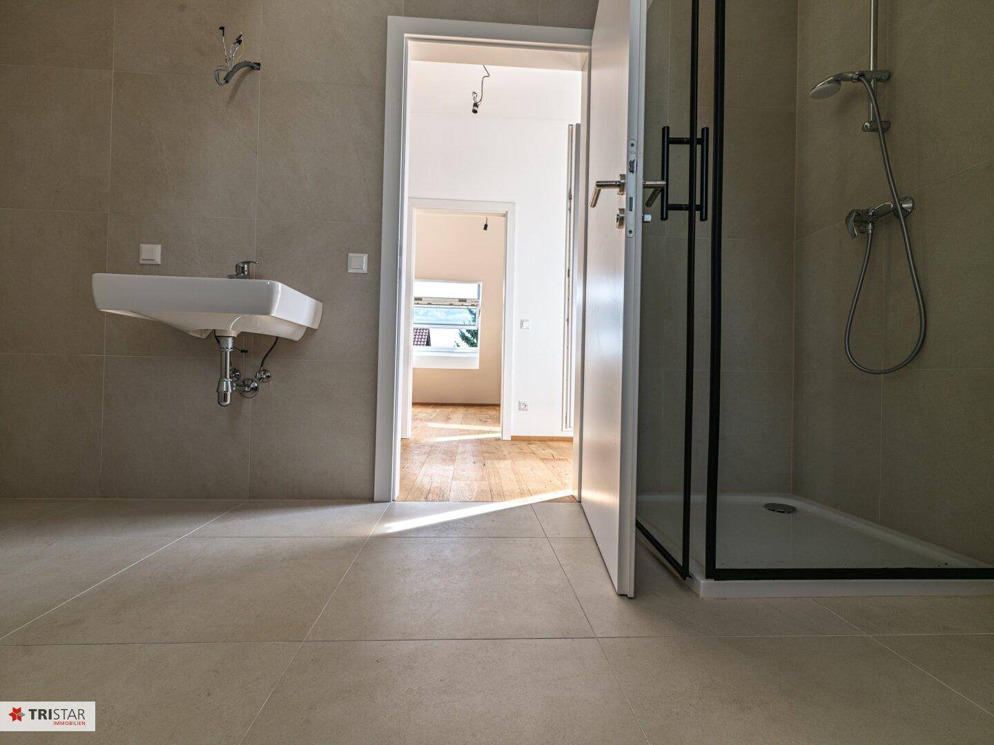 Helles Badezimmer mit Dusche und Waschbecken, mit Blick in den angrenzenden Flur.