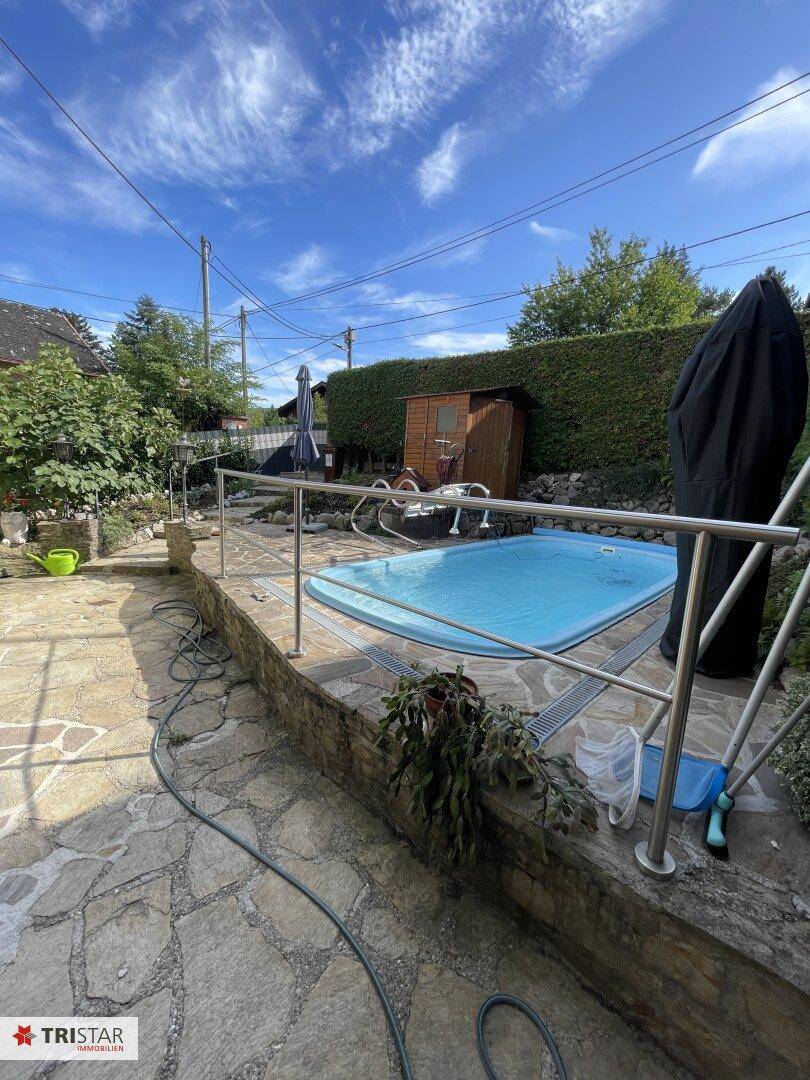 Außenbereich mit kleinem Pool, Steinterrasse und Metallgeländer, umgeben von grüner Bepflanzung.