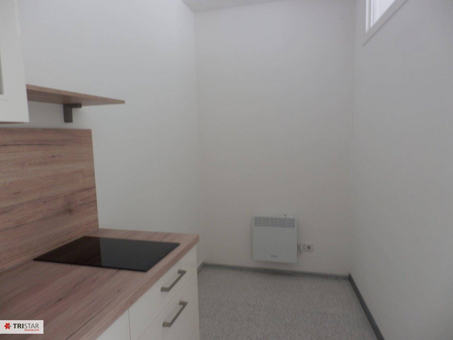 Kompakte Küchenzeile mit Kochfeld und Arbeitsplatte in einem kleinen, hellen Raum mit Heizkörper an der Wand.