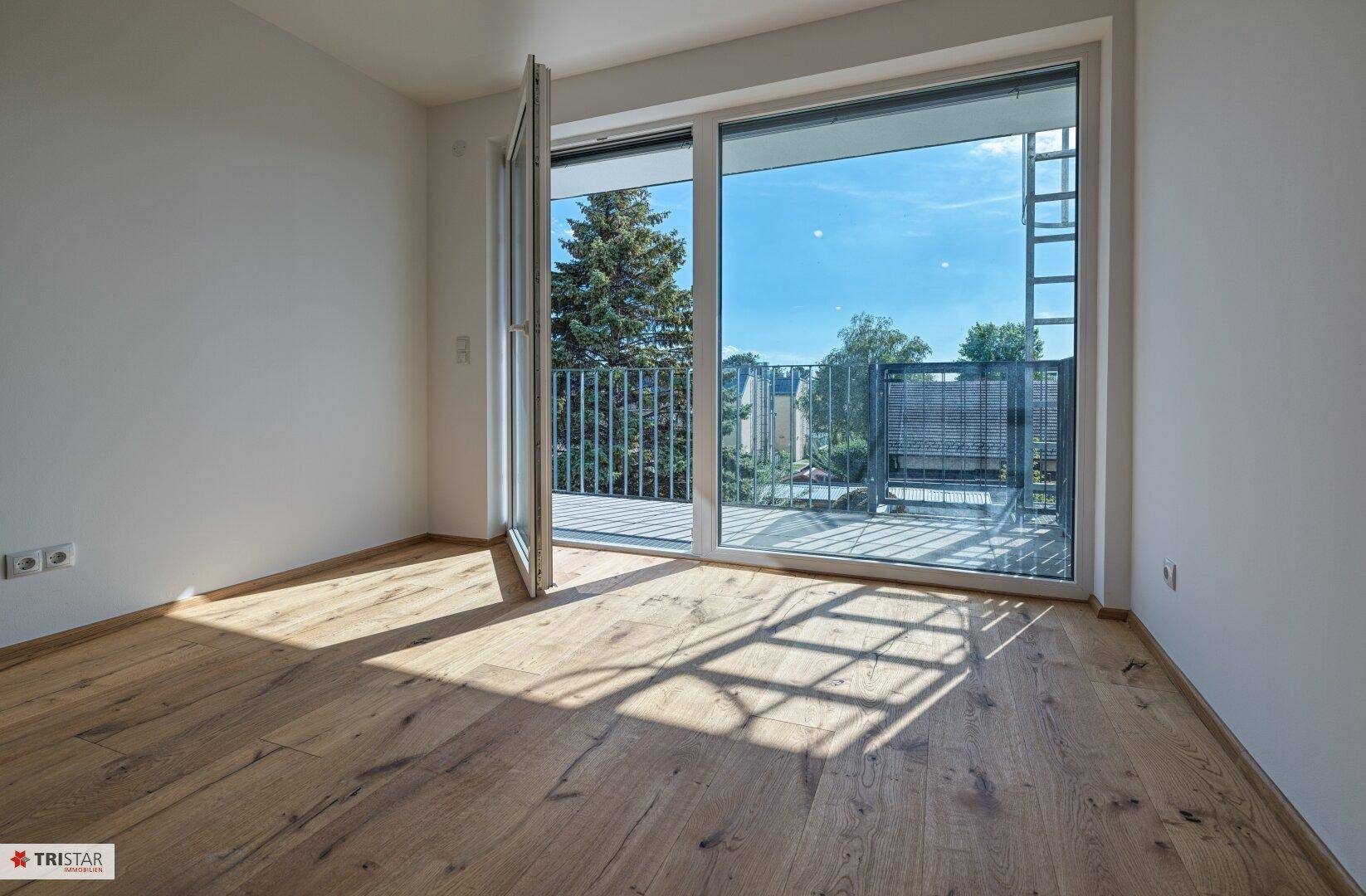 Heller Raum mit Eichenparkettboden und großer Schiebetür, die zu einem sonnigen Balkon führt.
