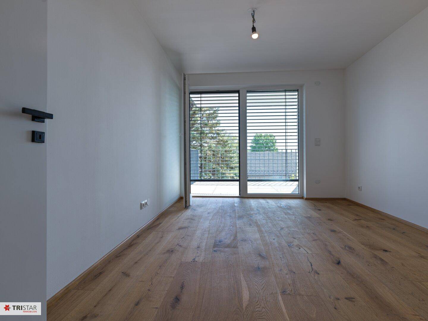 Helles Zimmer mit Holzboden und Balkonzugang, geschützt durch moderne Jalousien.