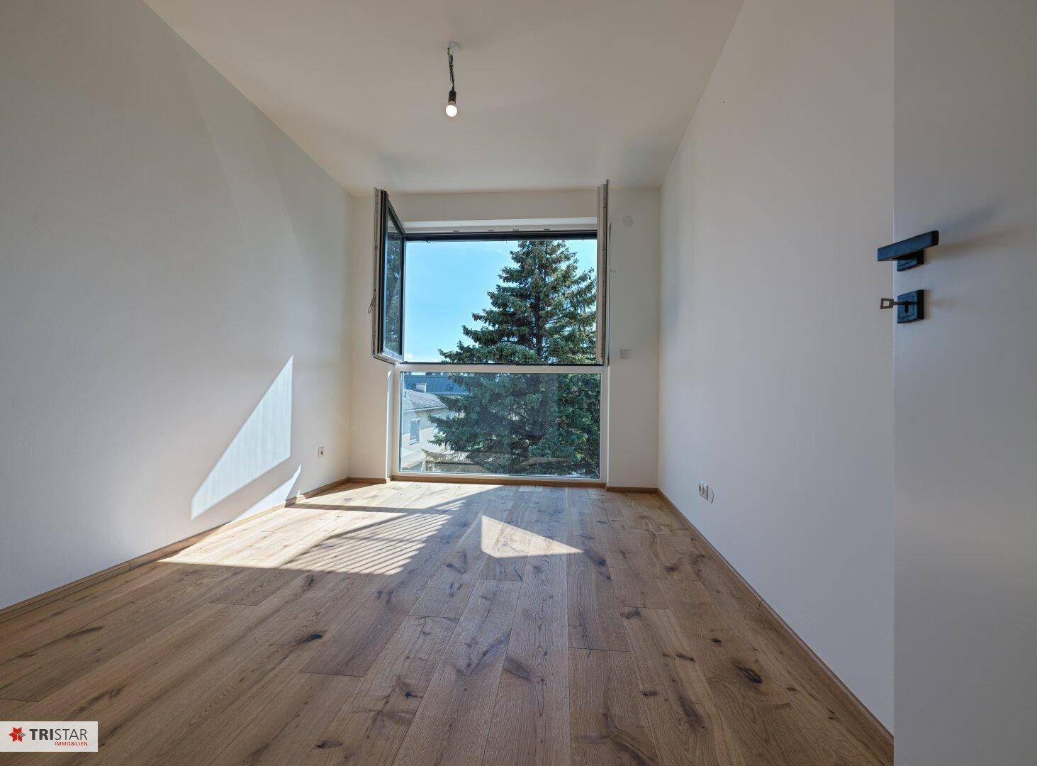 Helles Zimmer mit Holzboden und großem Fenster, das viel Tageslicht hereinlässt.