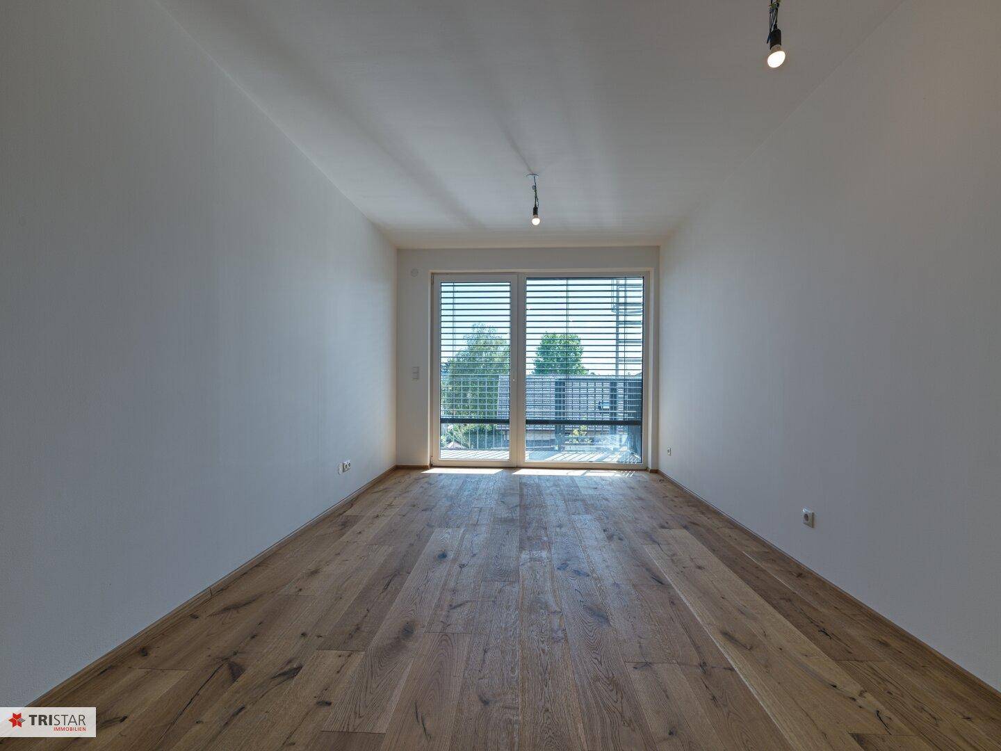 Helles Zimmer mit Holzboden und Zugang zu einem Balkon, ausgestattet mit Jalousien.
