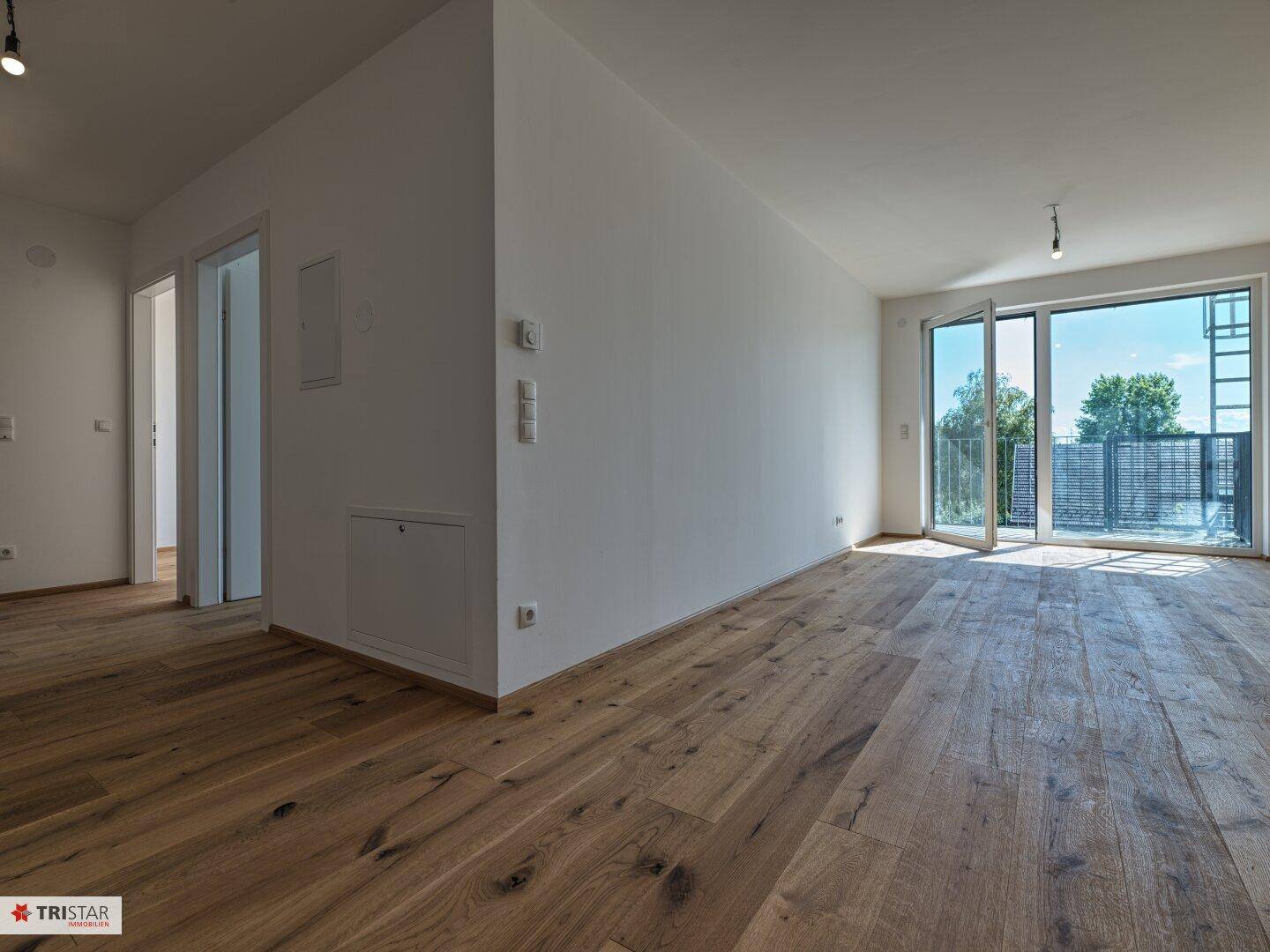 Geräumiger Wohnbereich mit Holzboden und großen Fenstern, die auf einen Balkon führen.