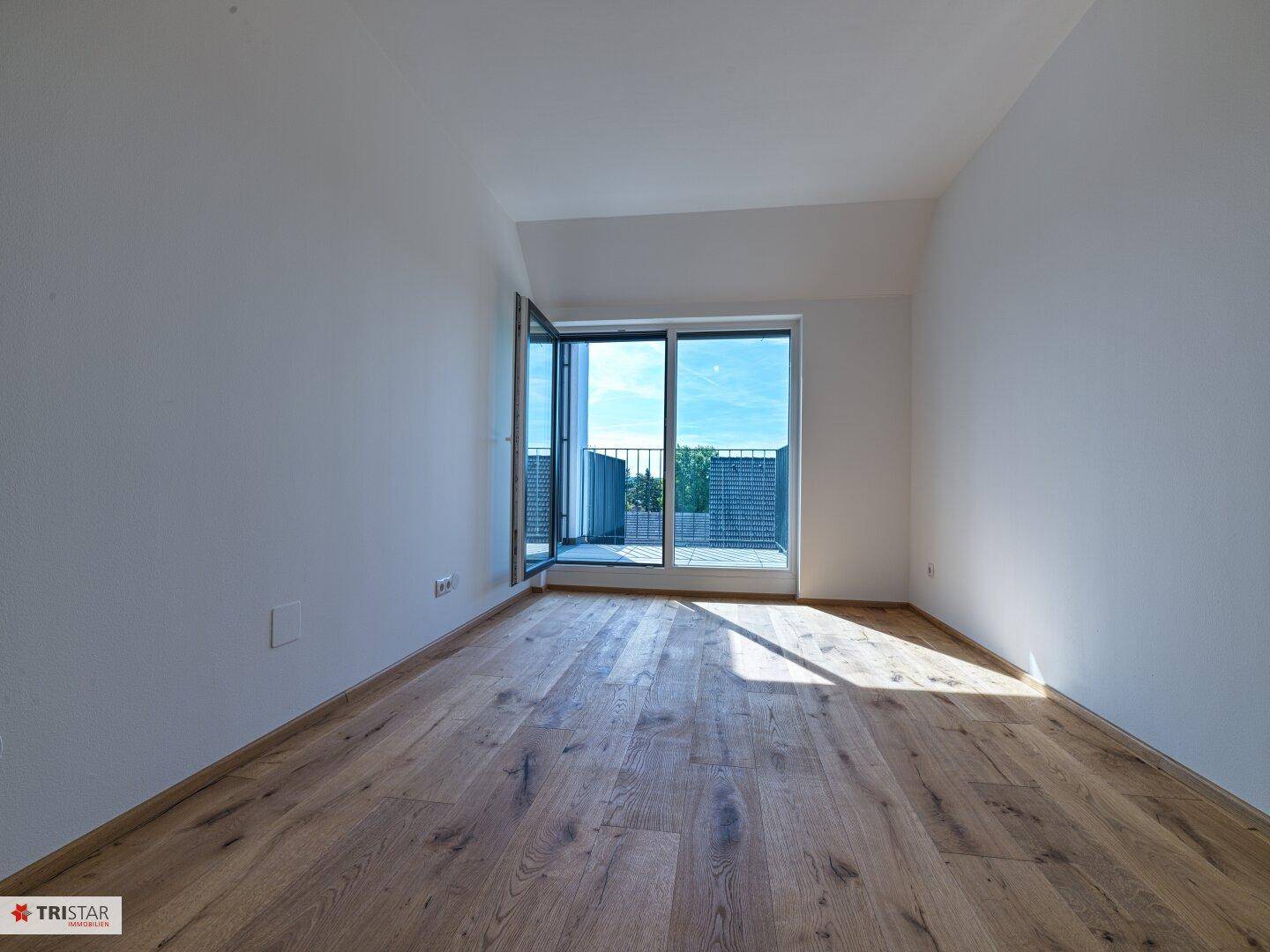 Heller Raum mit Holzboden und großem Fenster, das Zugang zu einem Außenbereich bietet.