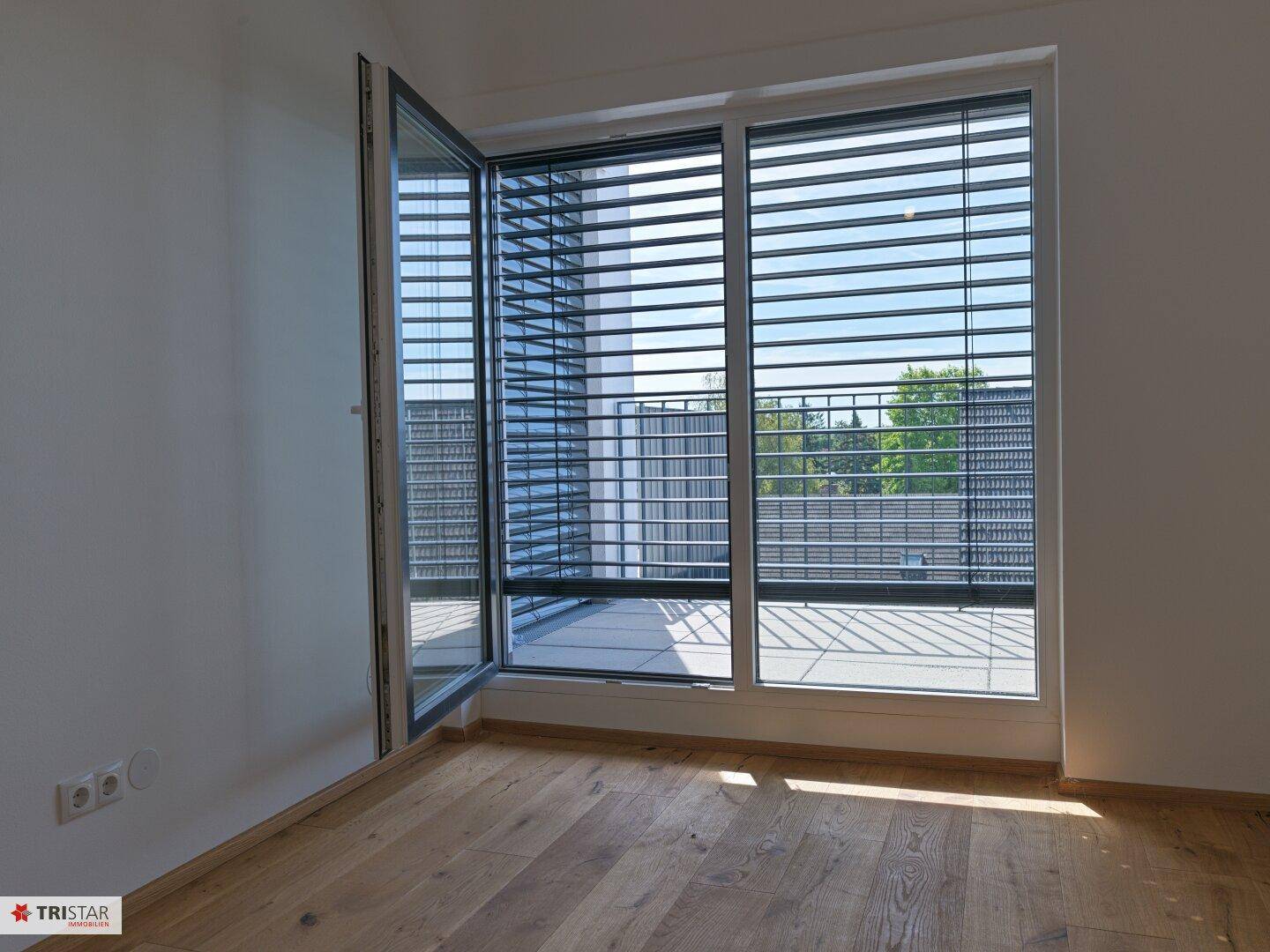 Sonniger Raum mit Holzboden und großen Fenstern mit Jalousien, ideal als Schlafzimmer.