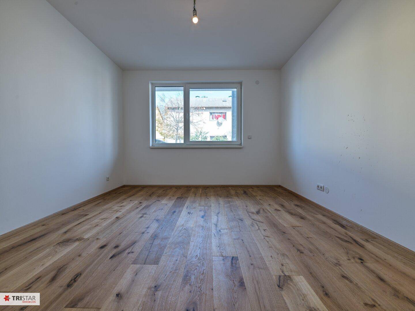 Heller, leerer Raum mit Holzboden und einem großen Fenster, das viel Tageslicht hereinlässt.
