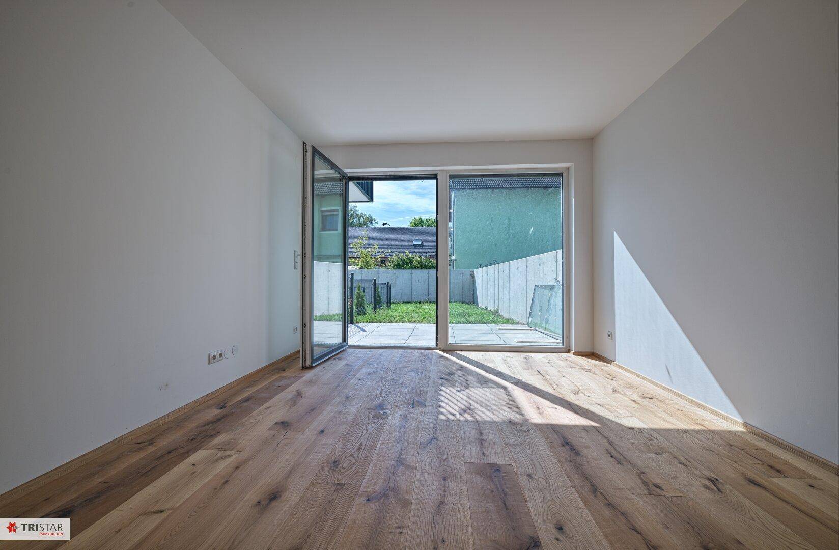 Heller, leerer Raum mit Holzboden und großen Glasschiebetüren, die zu einer Terrasse und dem Garten führen.