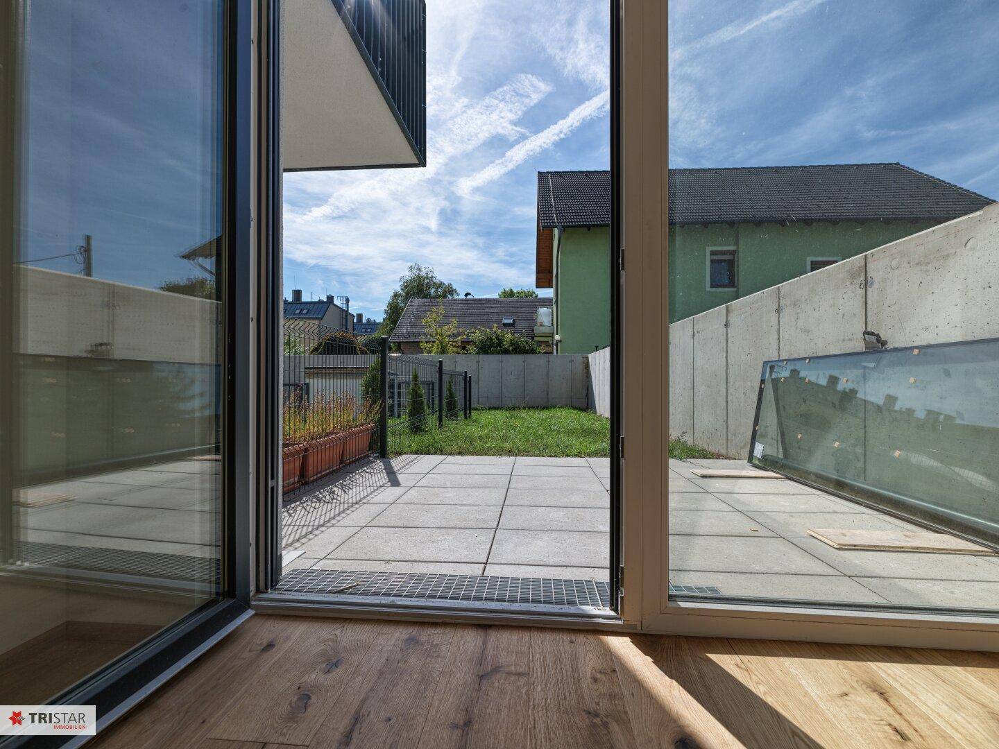 Blick aus einem hellen Innenraum mit Holzboden auf eine geflieste Terrasse und einen kleinen Gartenbereich.