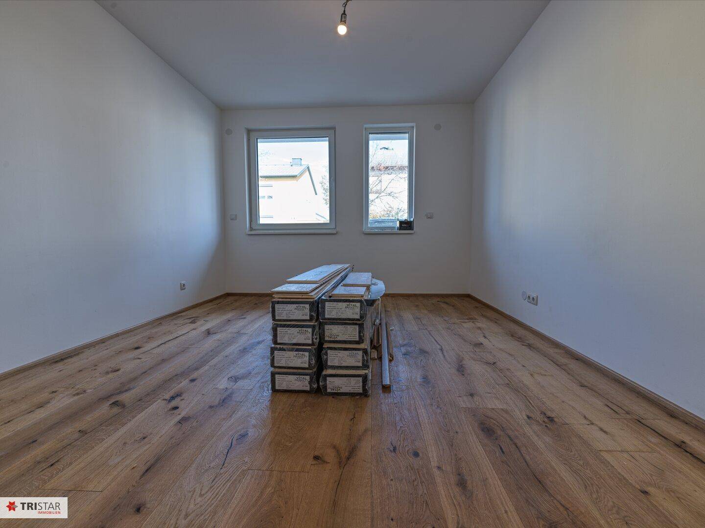 Helles Zimmer mit Holzboden und zwei Fenstern, in dem Baumaterialien gelagert sind.