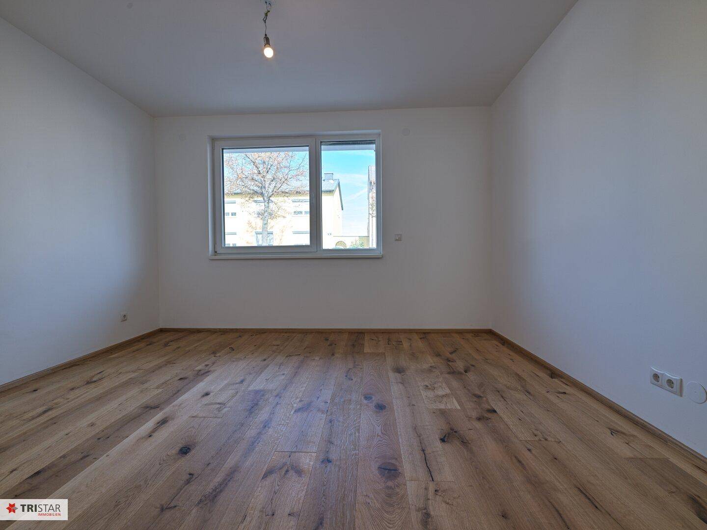 Leeres Zimmer mit Holzboden und einem großen Fenster, das viel Tageslicht bietet.