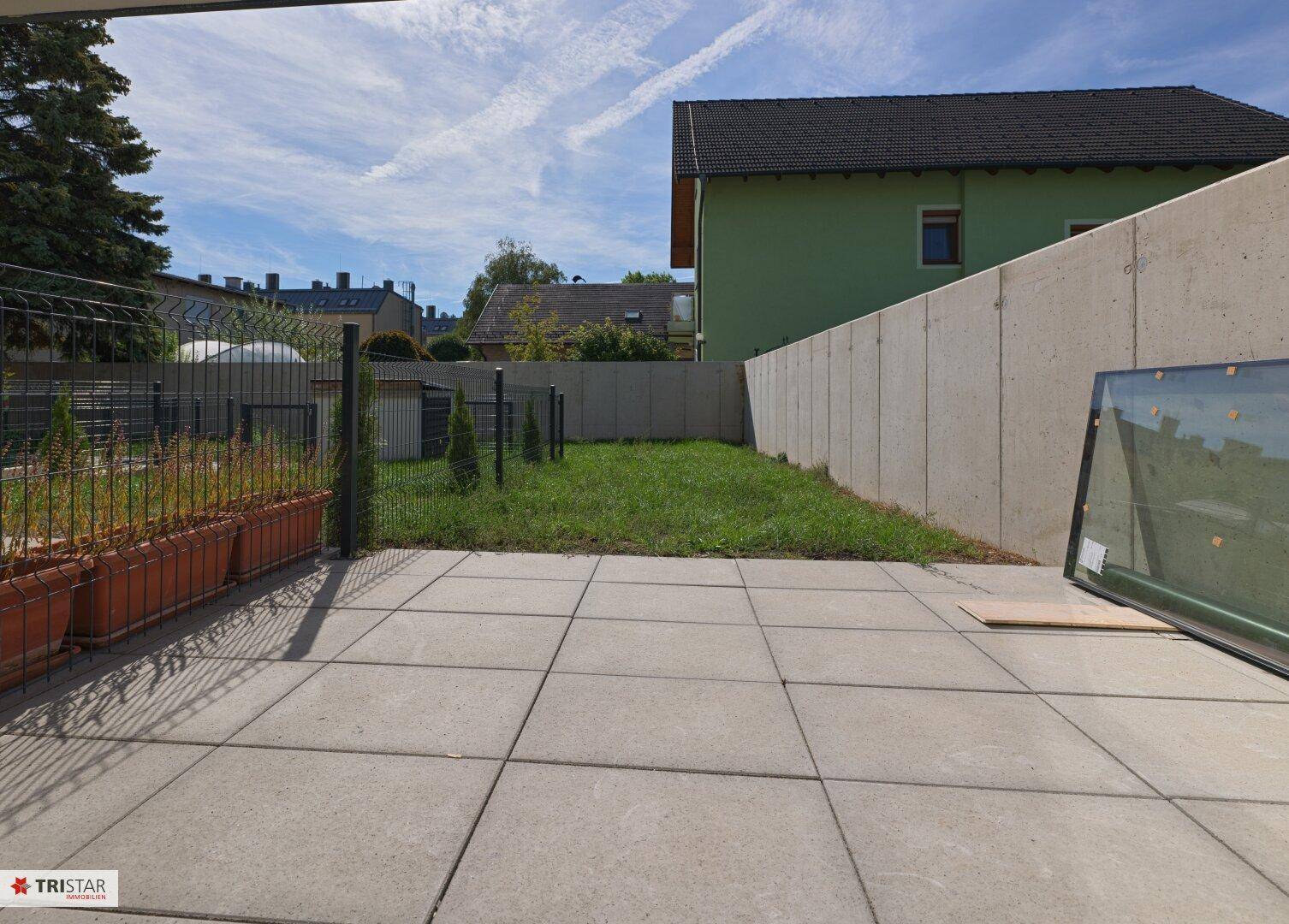 Gepflasterte Terrasse mit angrenzendem Rasenbereich und einer Betonmauer unter blauem Himmel.