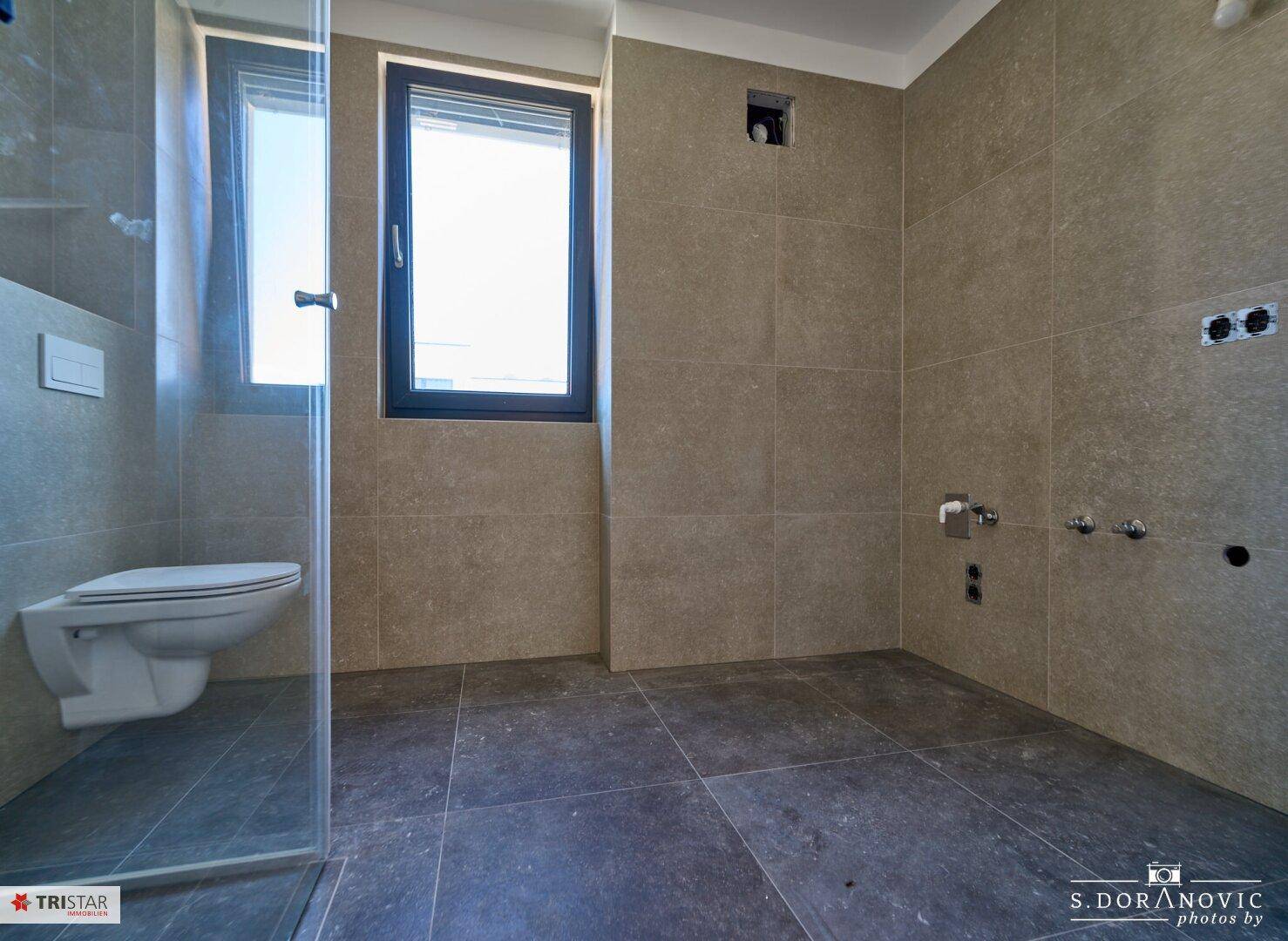 Modernes Badezimmer mit grauen Fliesen, wandhängendem WC und vorbereitetem Duschbereich.