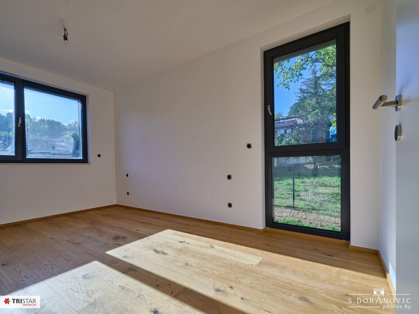 Helles Schlafzimmer mit Holzboden und großem Fenster mit Blick auf die grüne Umgebung.