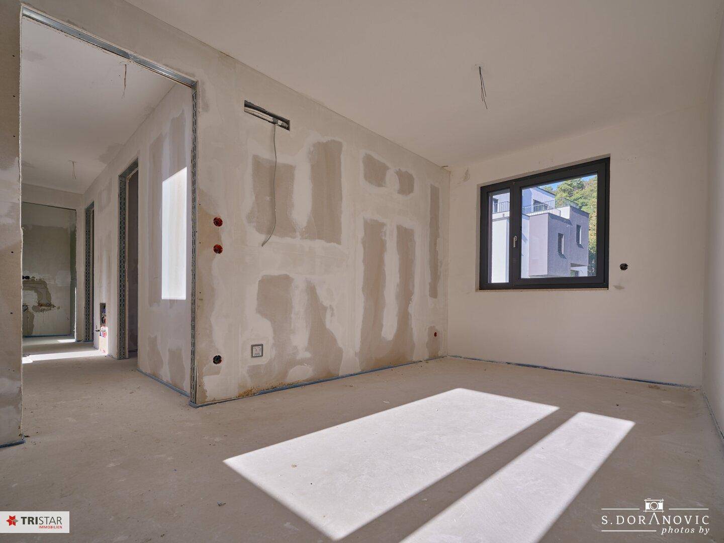 Unfertiger Innenraum mit Betonboden, Sonnenlicht und Blick auf ein modernes Gebäude durch das Fenster.