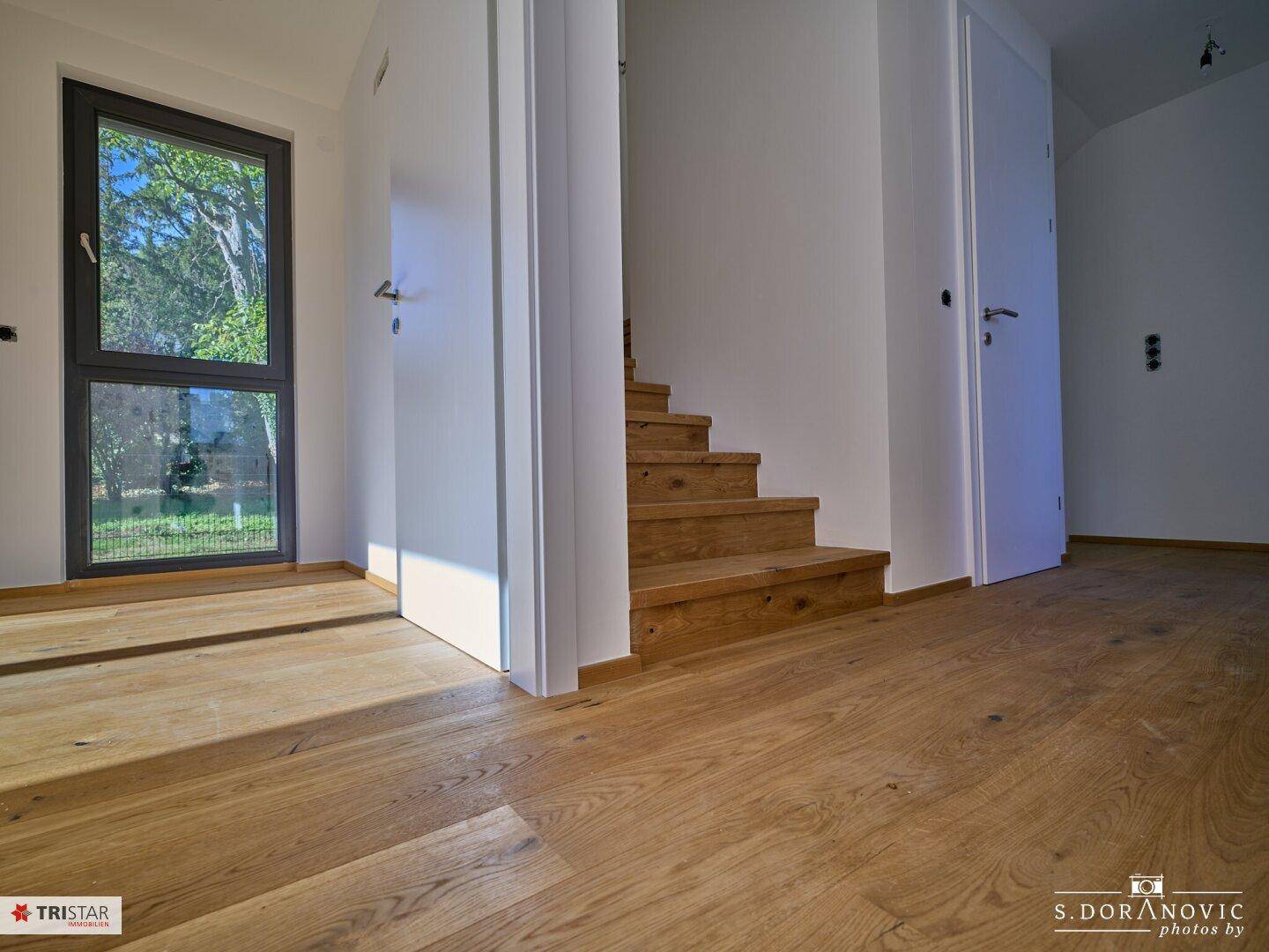 Heller Flurbereich mit neuem Hartholzboden, Holztreppe und einer hohen Fenstertür mit Zugang nach draußen.