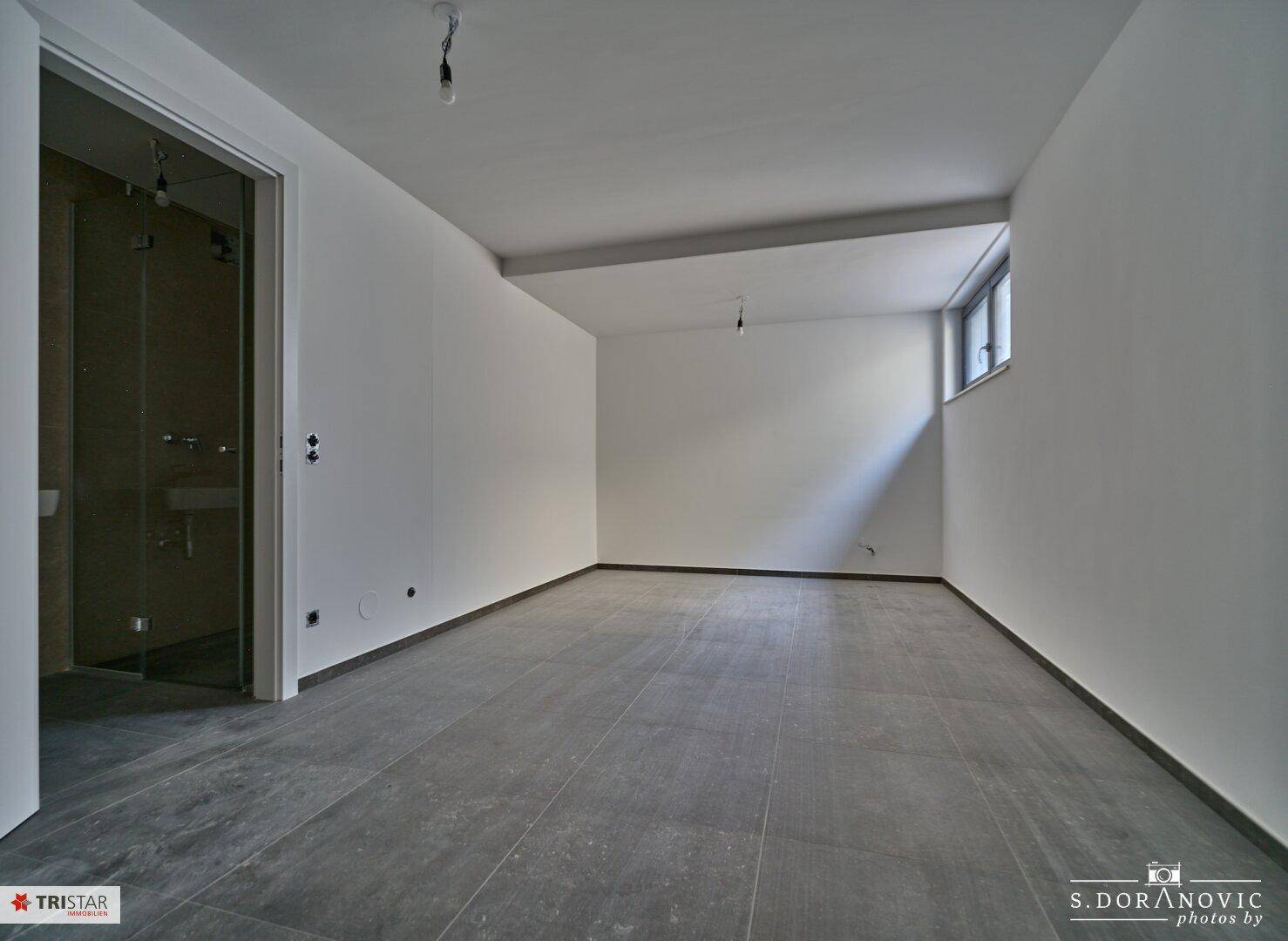 Kellerraum mit grauen Fliesen, weißen Wänden und Zugang zu einem Badezimmer, mit kleinem Fenster.