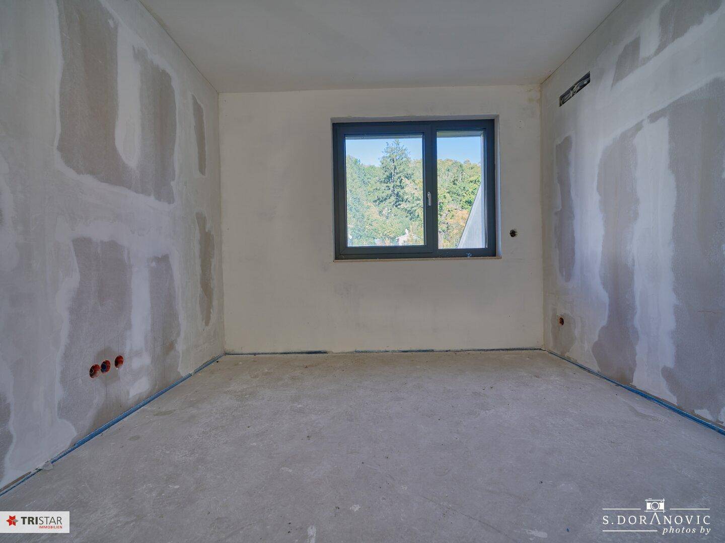 Unfertiger Raum mit Betonboden und Fenster, das einen Blick ins Grüne ermöglicht.