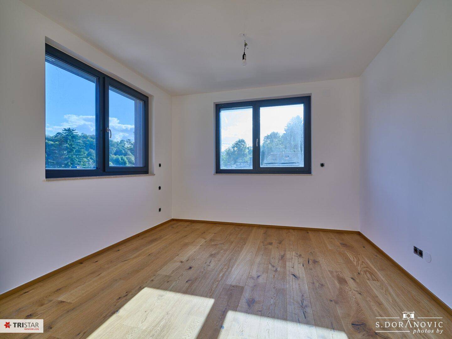 Helles Zimmer mit Holzboden und großen Fenstern, die einen Blick ins Grüne bieten.