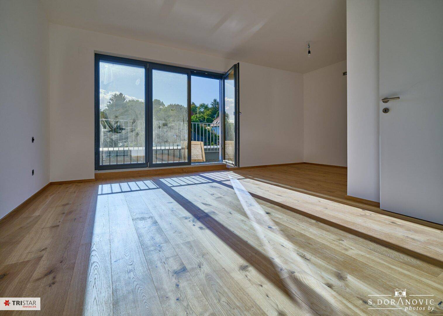 Geräumiger Raum mit Holzboden und großer Glasfront, die Zugang zu einem Balkon bietet.