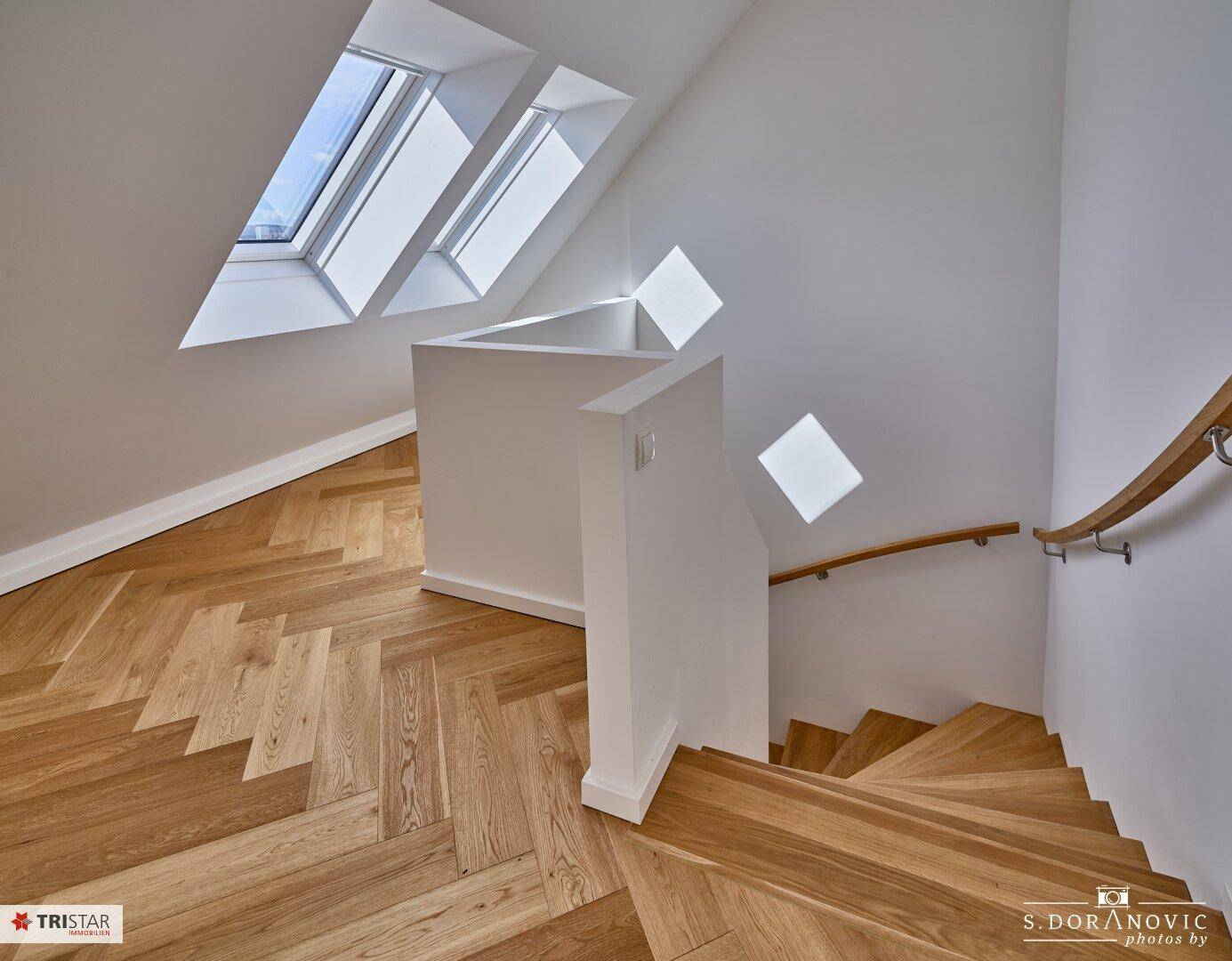 Treppenaufgang mit Holzstufen und Handlauf, führt zu einem hellen Dachgeschossbereich.