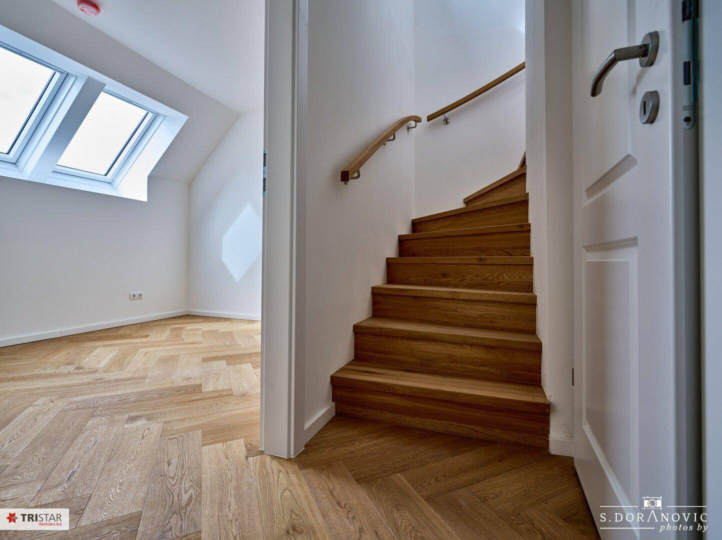 Treppenaufgang mit Holzstufen und Handlauf, führt zu einem hellen Zimmer mit Dachfenstern.