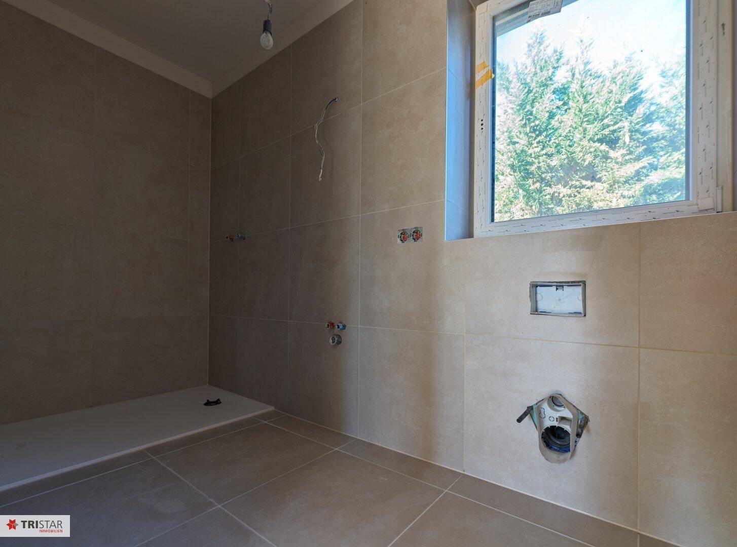 Unfertiges Badezimmer mit beigefarbenen Fliesen, Duschbereich und Fenster, bereit für die Installation.