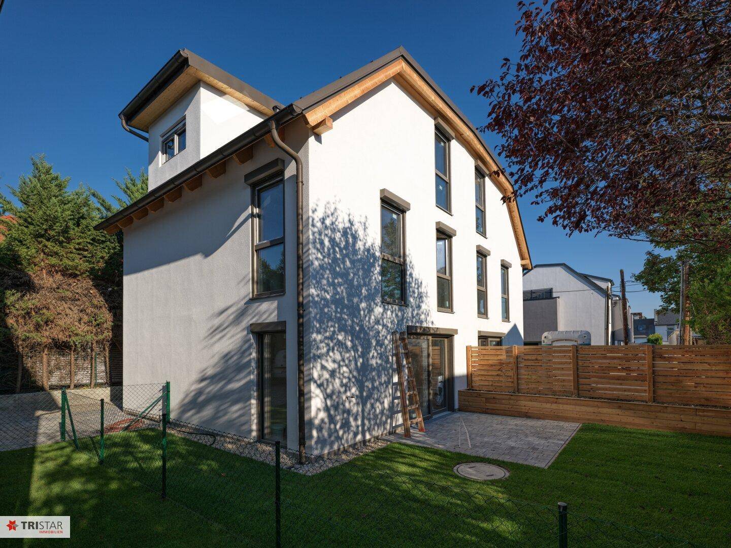 Rückansicht des modernen Hauses mit Terrasse und weitläufigem Rasenbereich unter blauem Himmel.