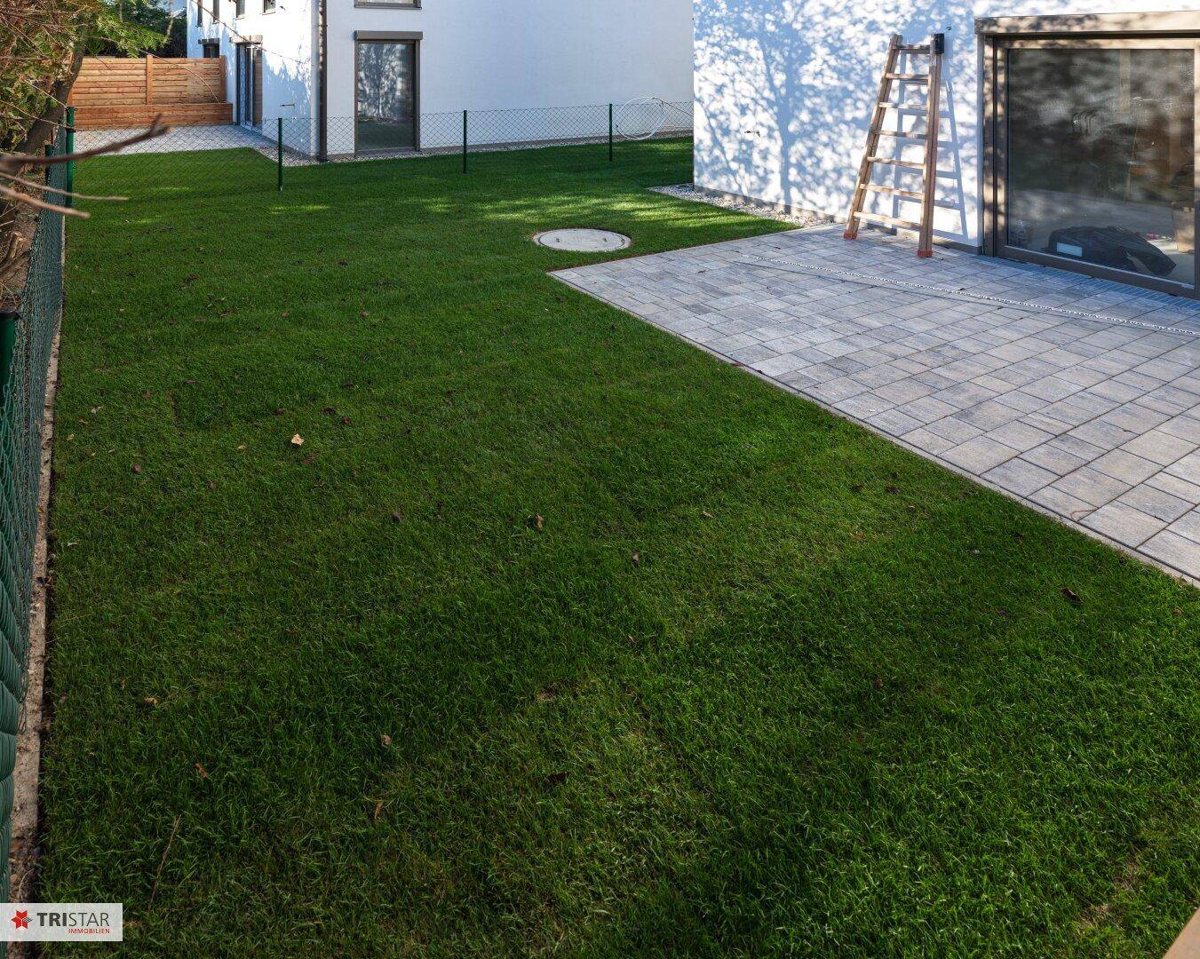 Frisch angelegter Rasenbereich neben einer gepflasterten Terrasse am Haus unter blauem Himmel.