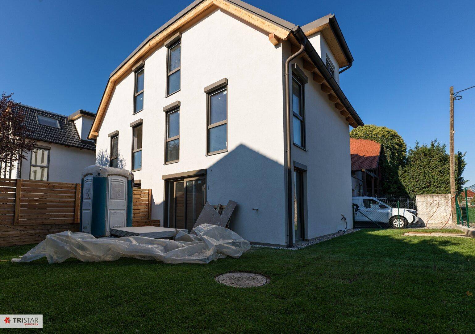 Seitenansicht des Hauses mit weißer Fassade, Rasenfläche und Bauutensilien im Vordergrund.