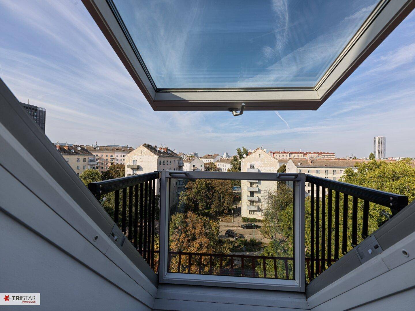 Aussicht aus einem Dachfenster über die Stadt mit grünen Bäumen und umliegenden Gebäuden.
