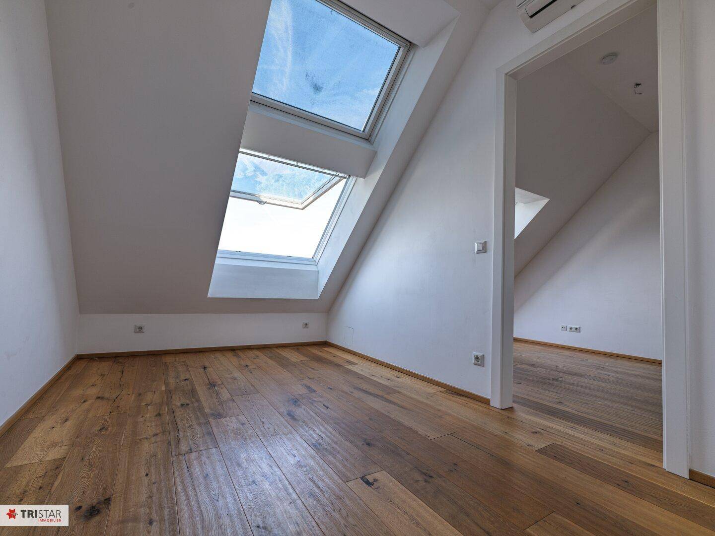 Geräumiger Dachgeschossraum mit Holzboden, schrägen Wänden und zwei großen Dachfenstern.