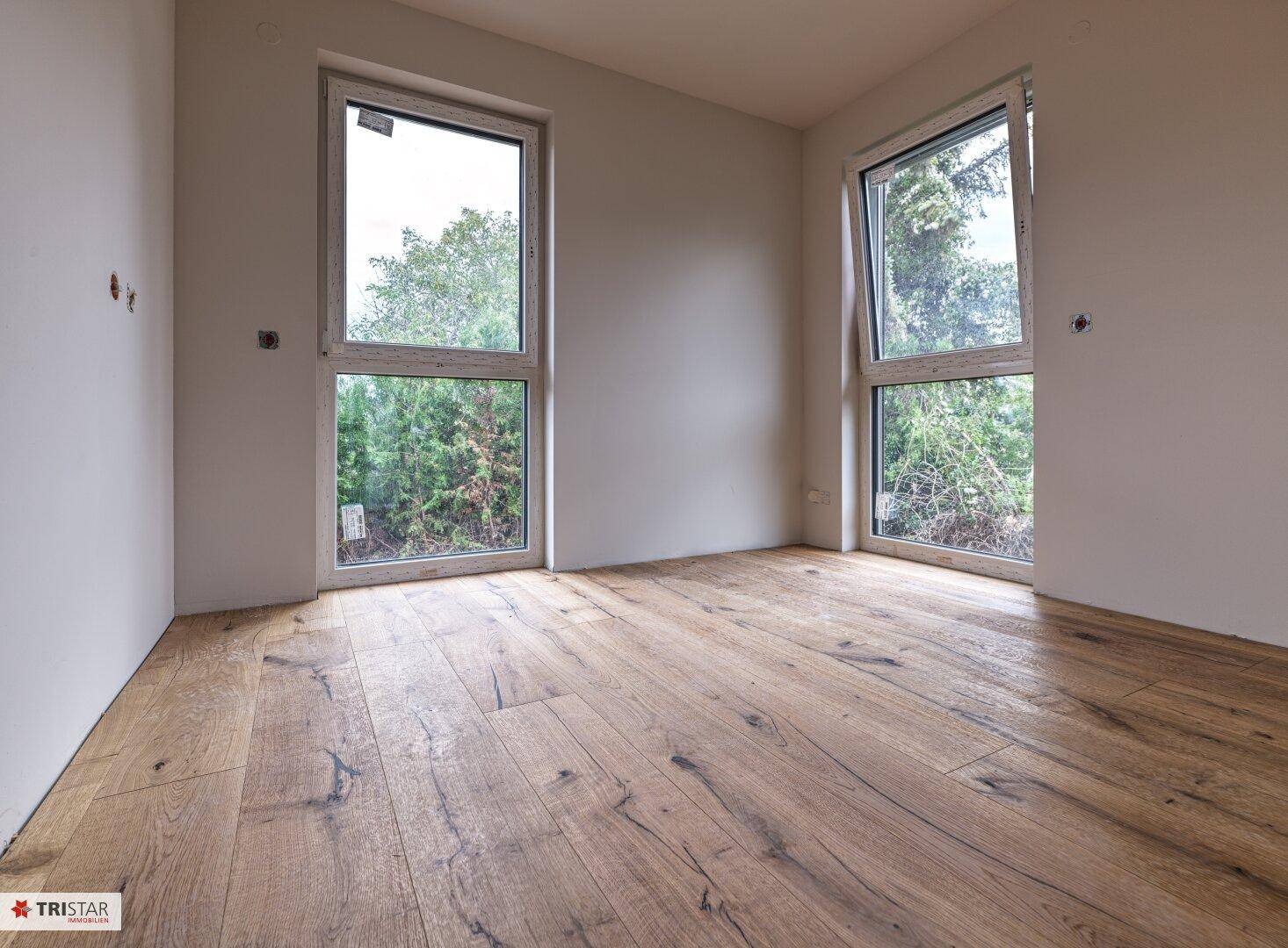 Leeres Zimmer im Bau mit Holzboden und zwei großen Fenstern mit Blick ins Grüne.