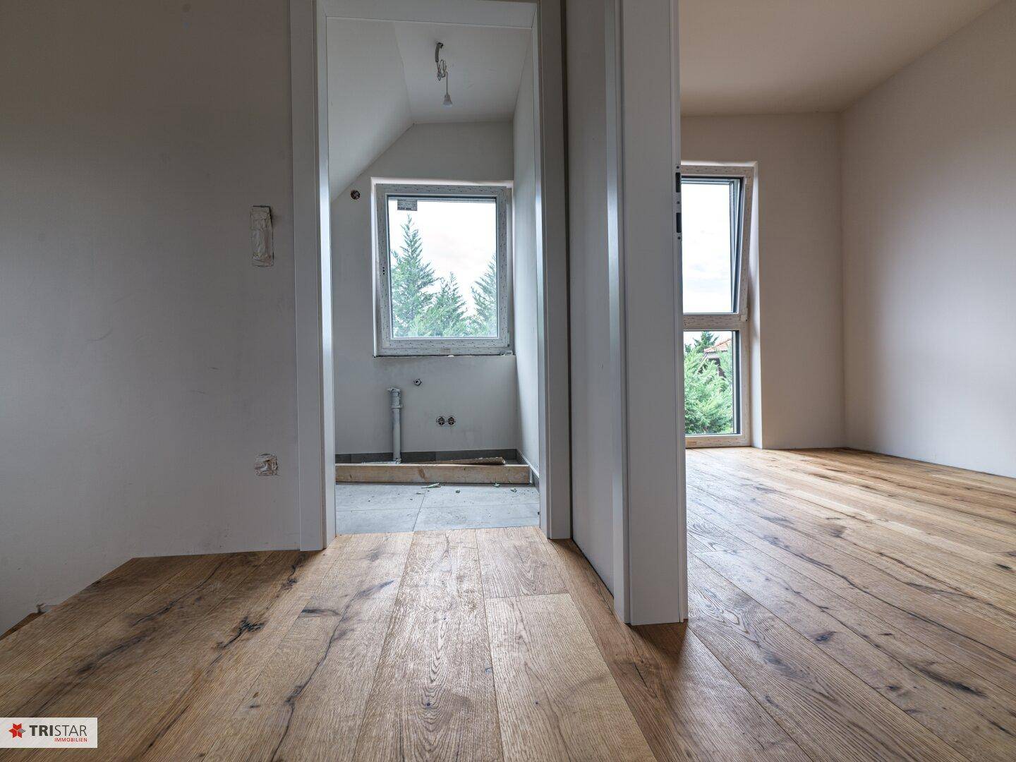 Blick durch einen Türrahmen auf zwei Räume, darunter ein Badezimmer mit Fenster und grauen Fliesen.
