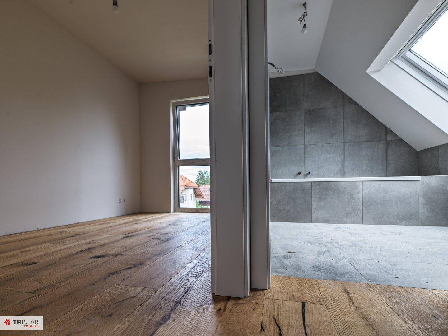 Blick durch einen Türrahmen auf zwei Räume, darunter ein Badezimmer mit Dachfenster und grauen Fliesen.