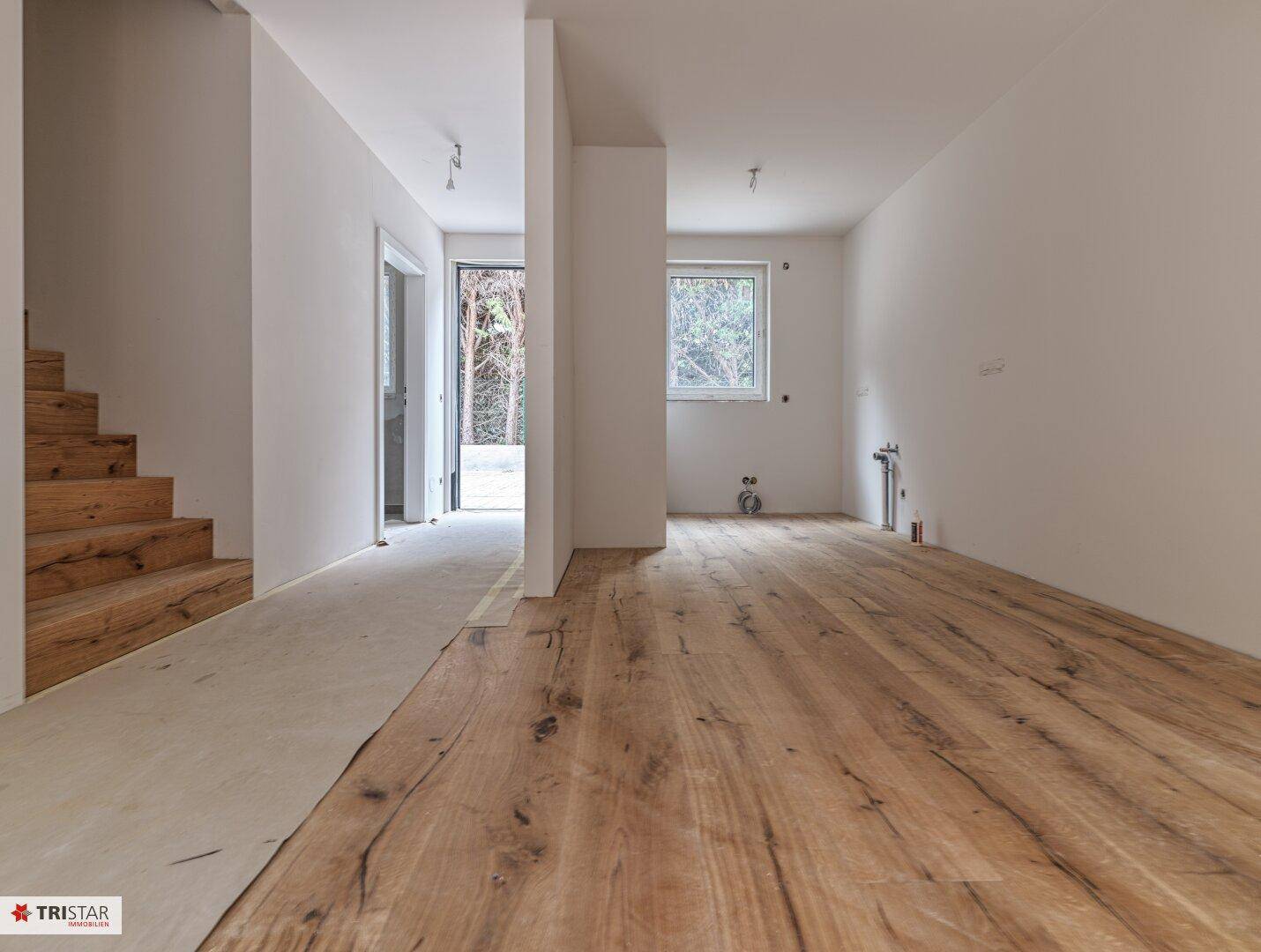 Offener Wohnbereich im Bau mit neuem Holzfußboden und Blick auf den Eingangsbereich und die Treppe.