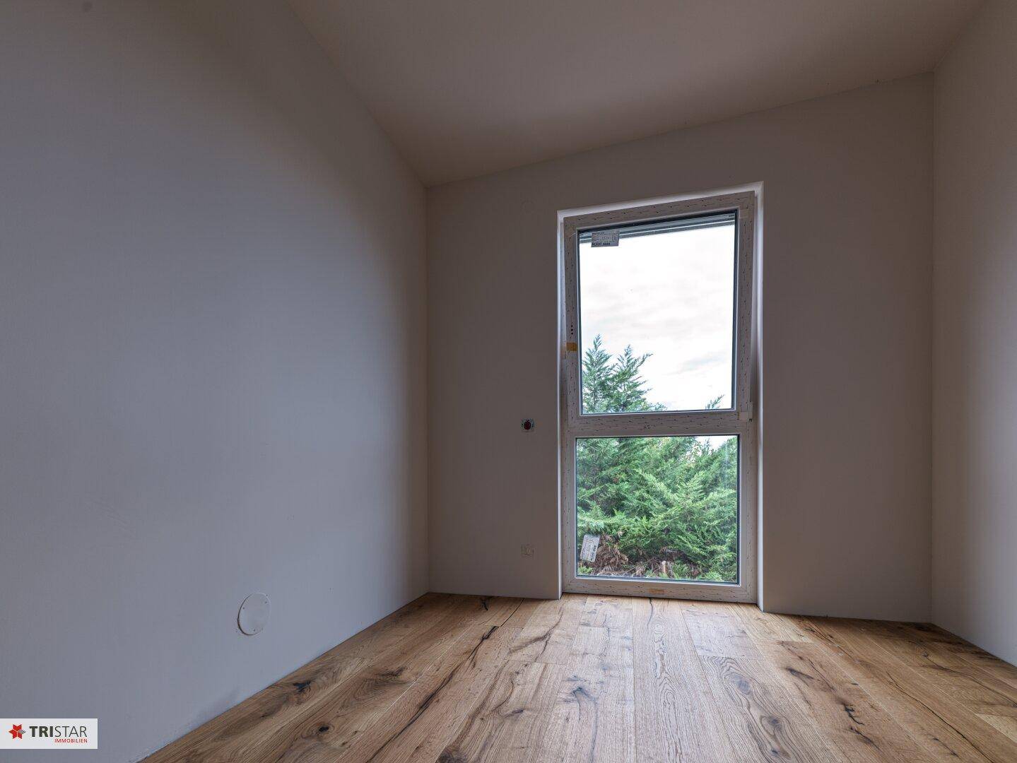 Unmöblierter Raum mit Holzdielenboden und einem hohen Fenster mit Blick ins Grüne.