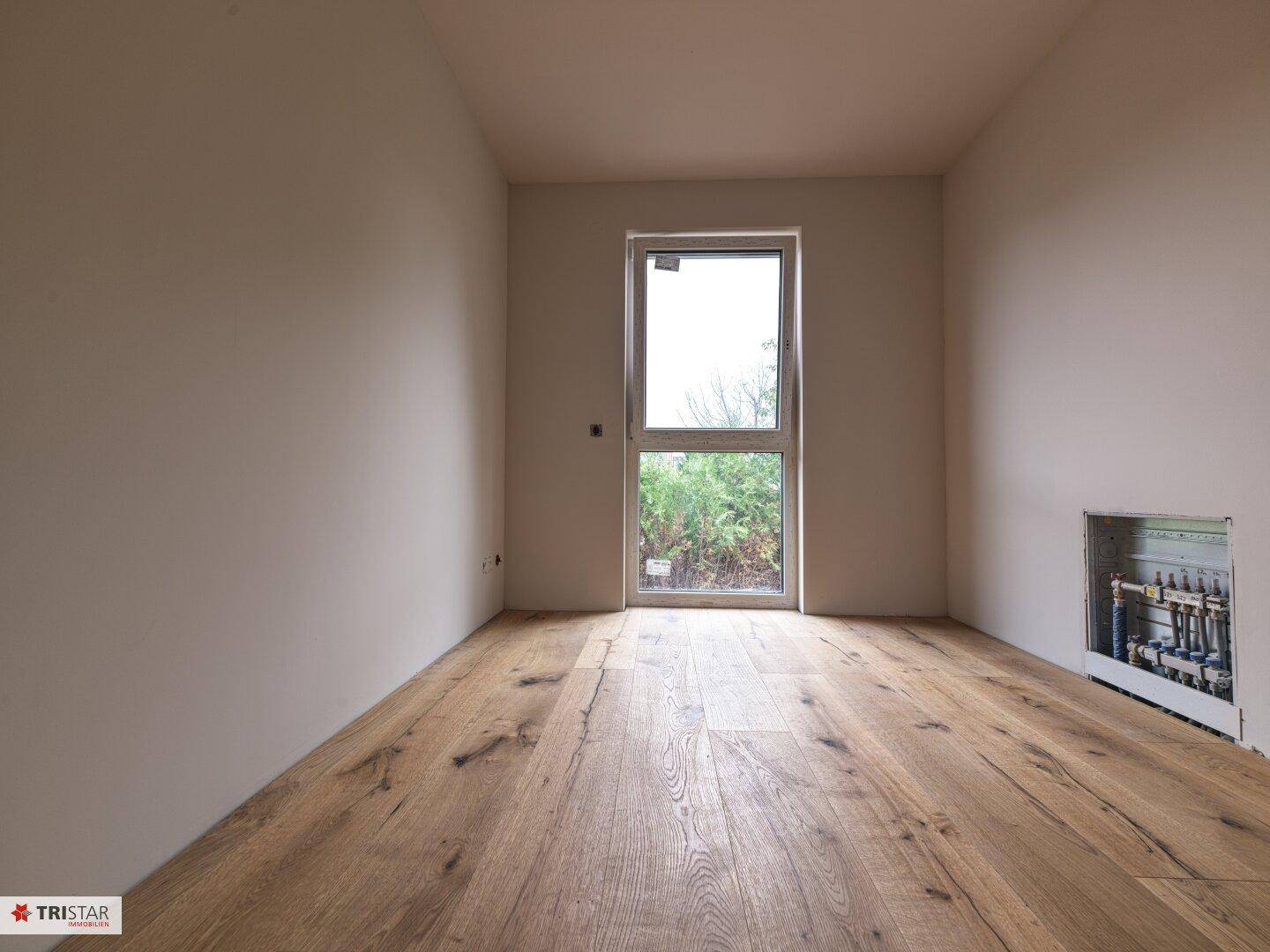 Unmöblierter Raum mit Holzdielenboden und einem hohen Fenster mit Blick ins Grüne.