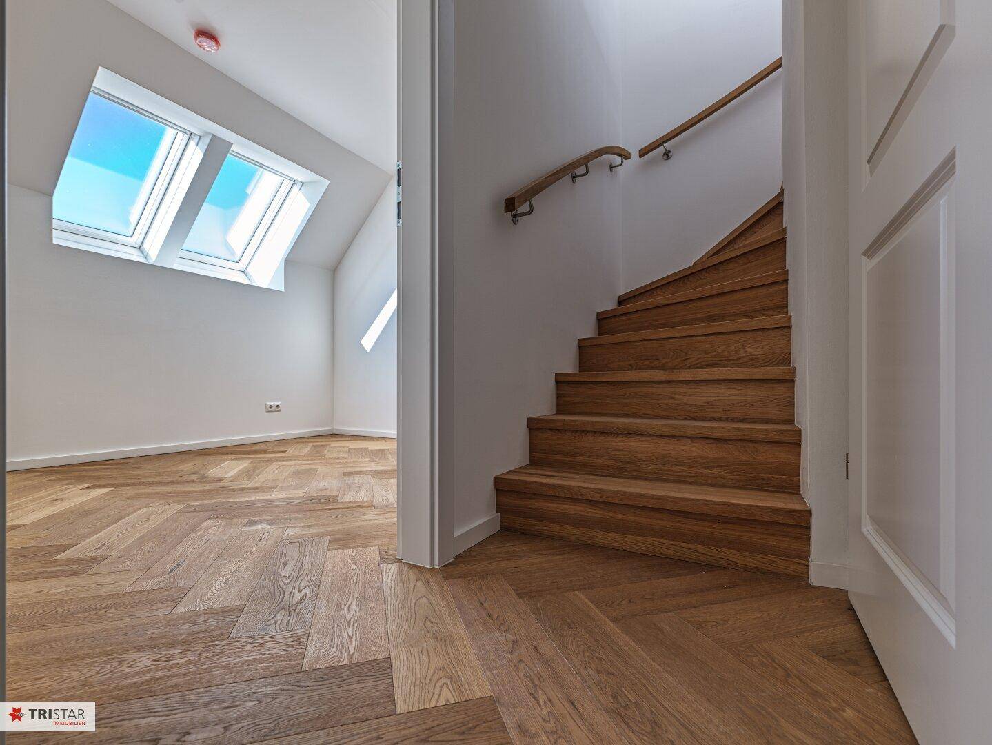 Blick von einem Raum auf eine elegante Holztreppe mit Handlauf und Dachfenster im Hintergrund.