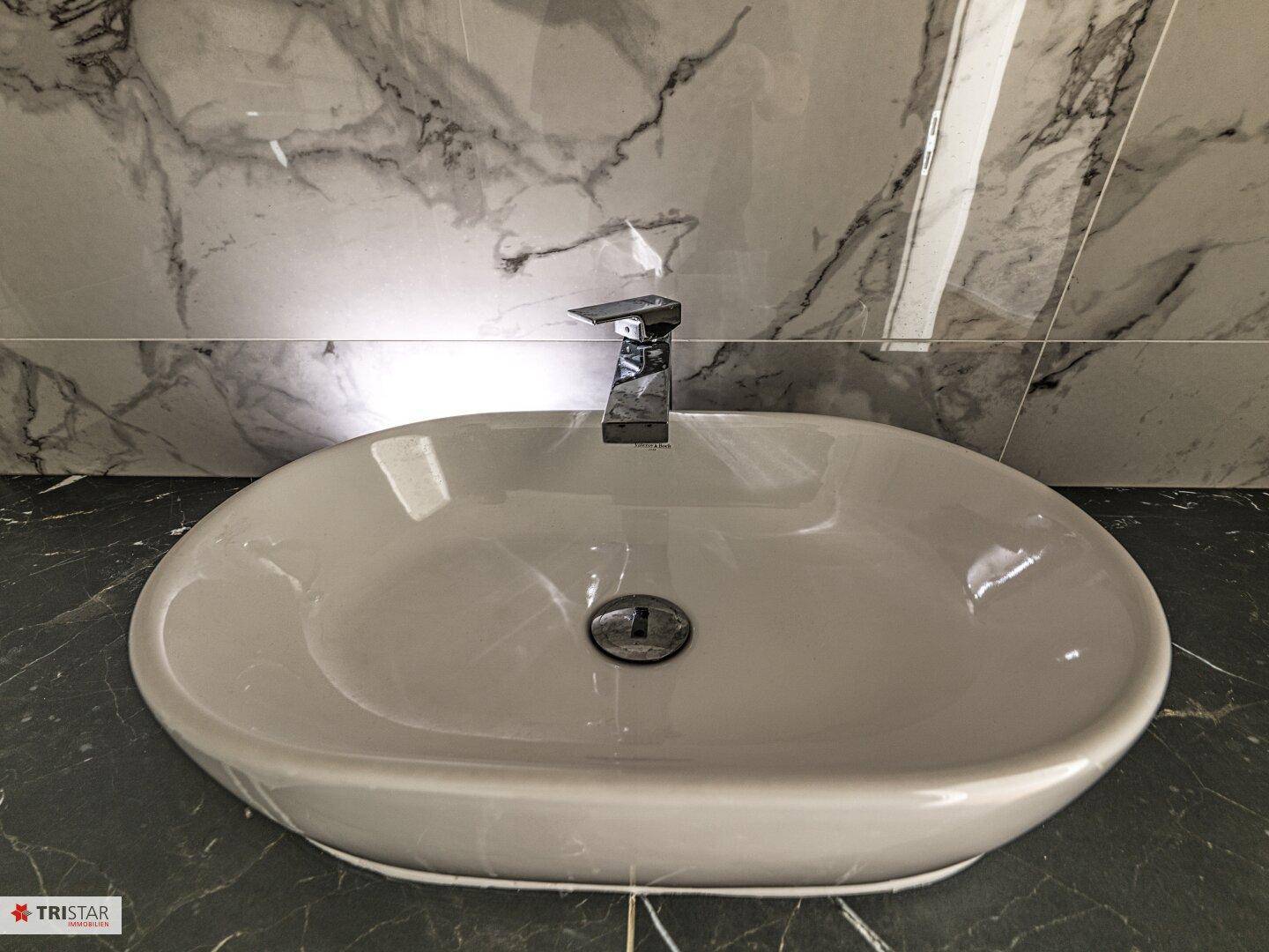 Modernes Waschbecken auf dunkler Arbeitsplatte mit Marmorfliesen an der Wand und Chromarmatur.