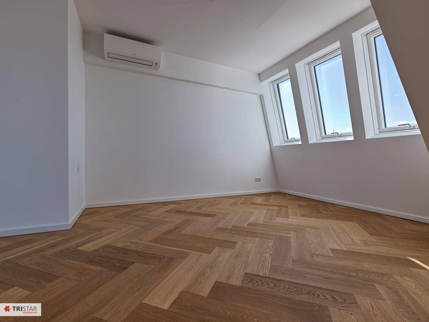 Heller Raum mit Fischgrätparkett, weißen Wänden, Fenstern und einer Klimaanlage an der Decke.