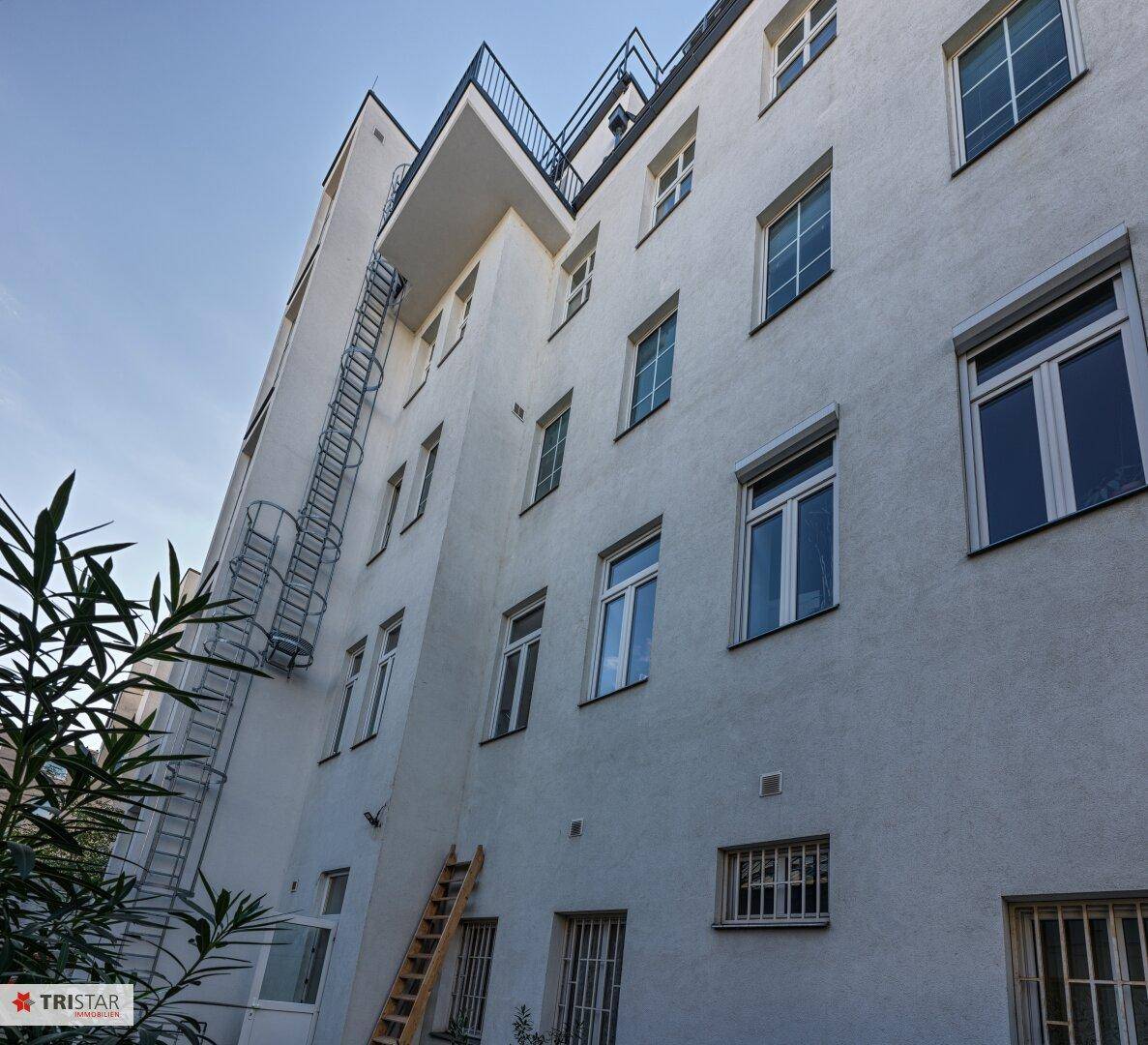 Seitenansicht eines Gebäudes mit mehreren Fenstern, einer Feuerleiter und einem kleinen Balkon.