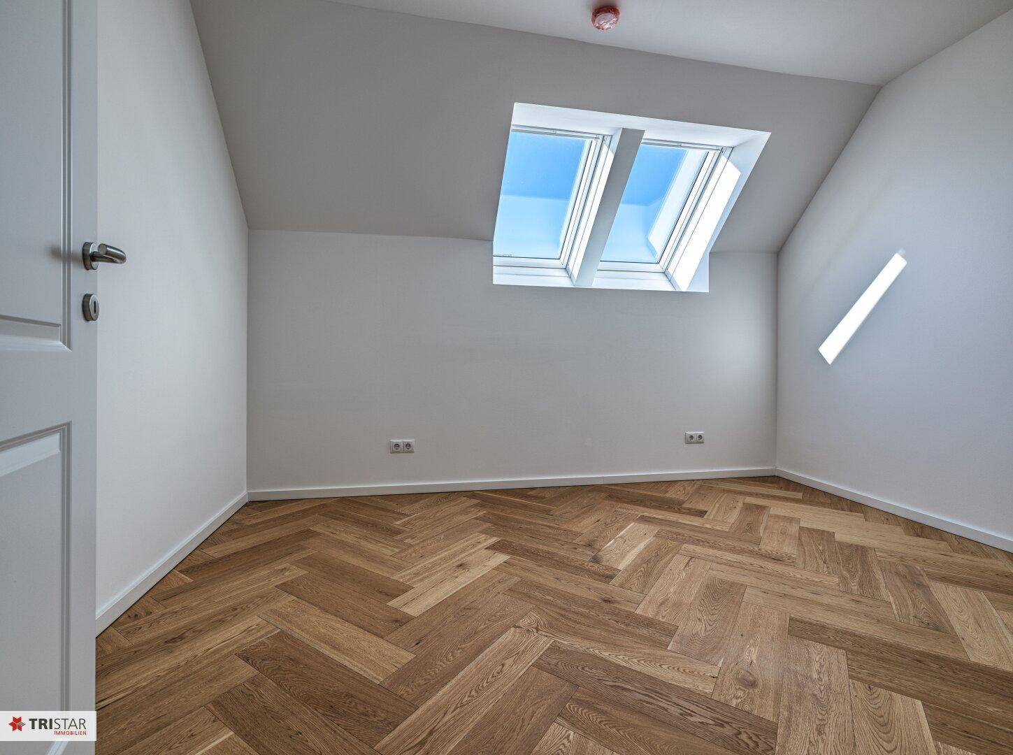 Helles Zimmer mit Dachschräge, Fischgrätparkett und zwei Dachfenstern, die den Raum erhellen.