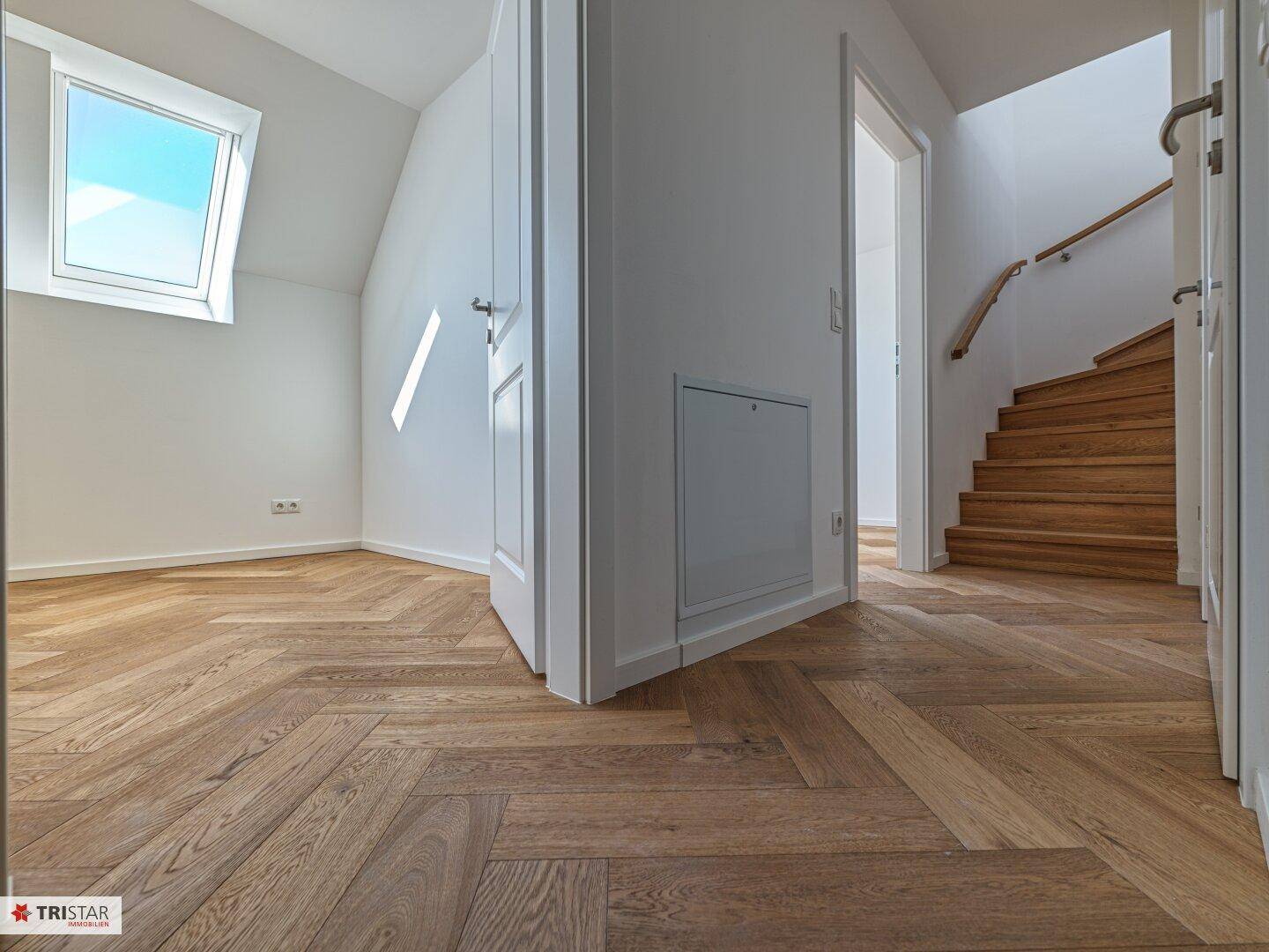 Heller Flur mit Fischgrätparkett, weißen Türen und einer Holztreppe, die nach oben führt.