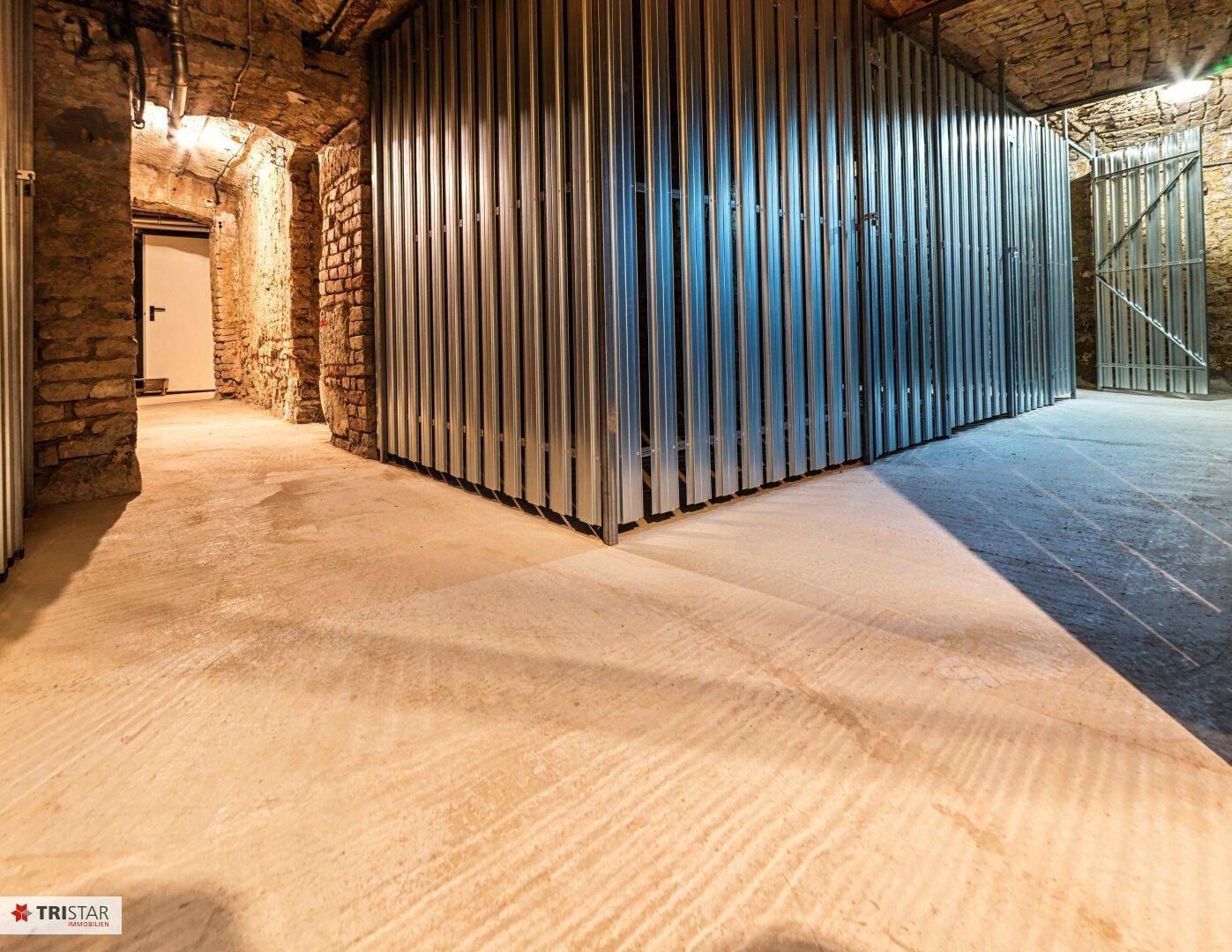 Historischer Kellerraum mit freiliegenden Ziegelwänden und modernen Metallabtrennungen für Abstellflächen.