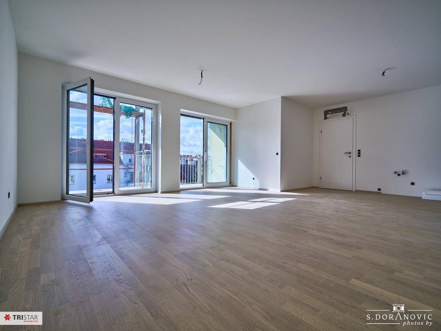 Weitläufiger Wohnbereich mit hellem Holzboden, großen Fenstern und Zugang zum Balkon.