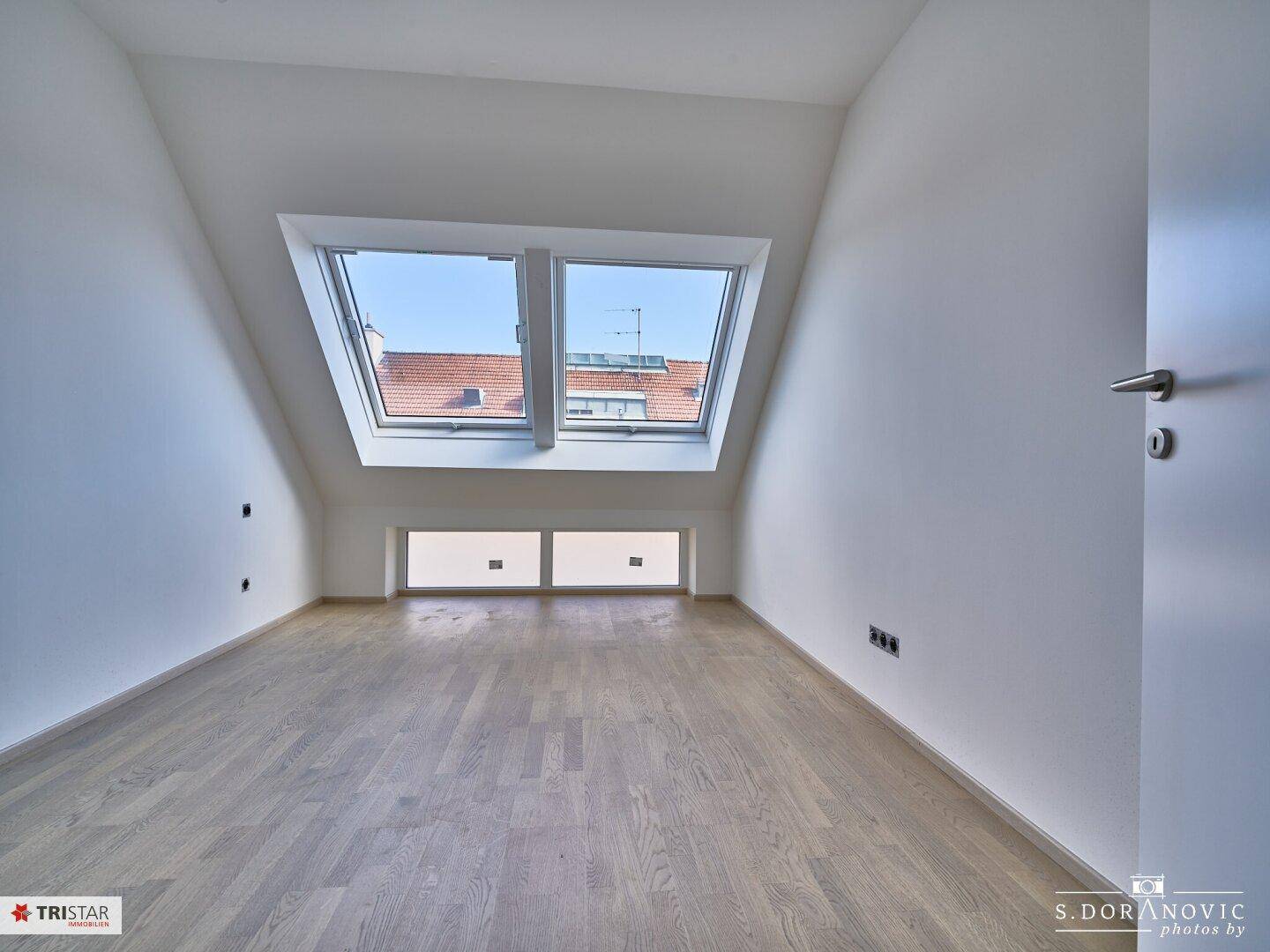 Helles Zimmer im Dachgeschoss mit schrägen Wänden, Holzboden und Dachfenstern.