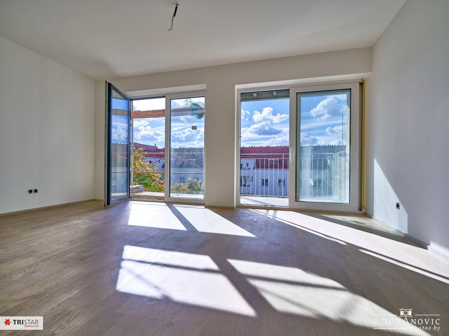 Heller, unmöblierter Raum mit Holzboden und großen Fenstern, die viel Tageslicht hereinlassen.