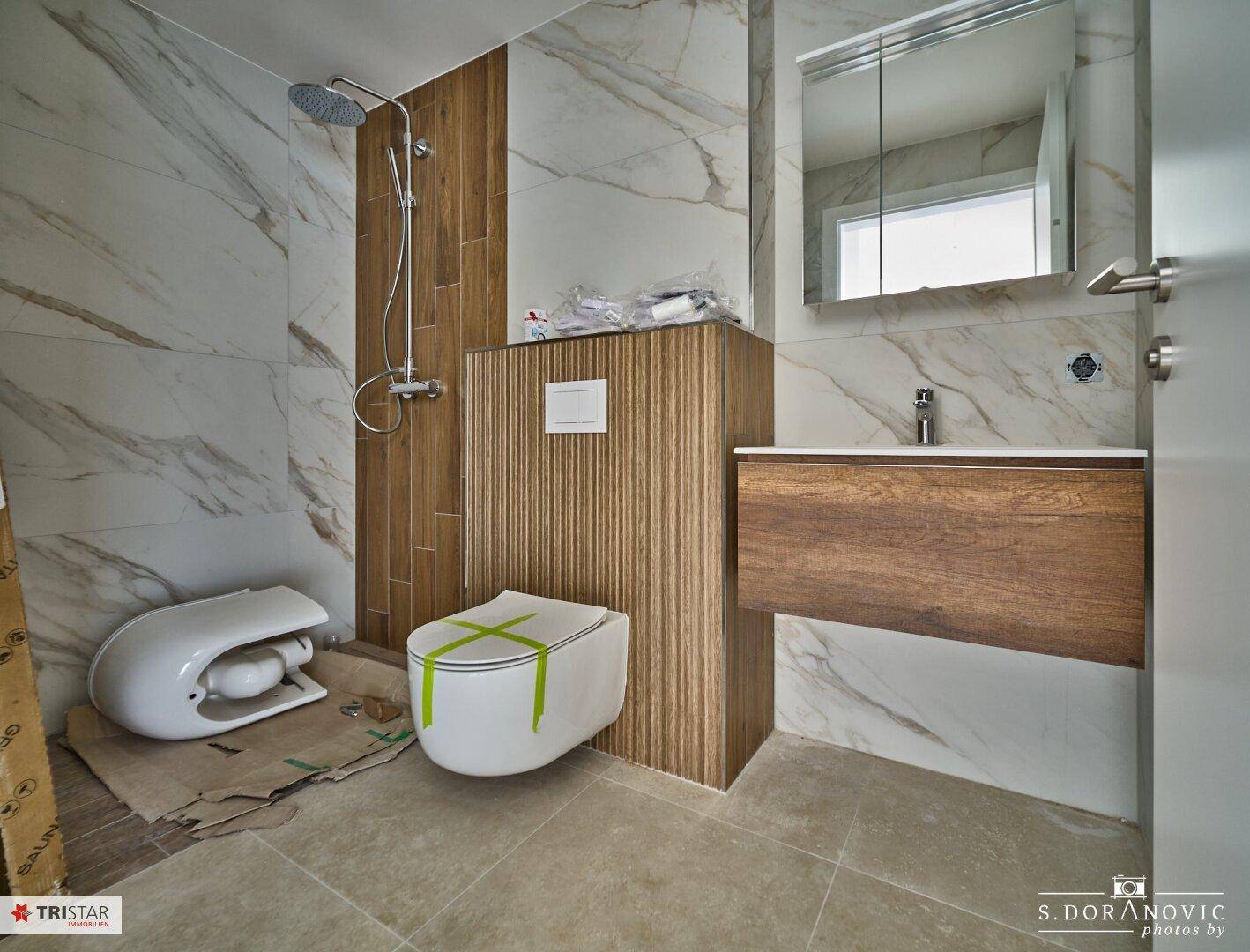 Modernes Badezimmer im Bau mit Wand-WC, Dusche, Waschtisch und Marmoroptik-Fliesen.