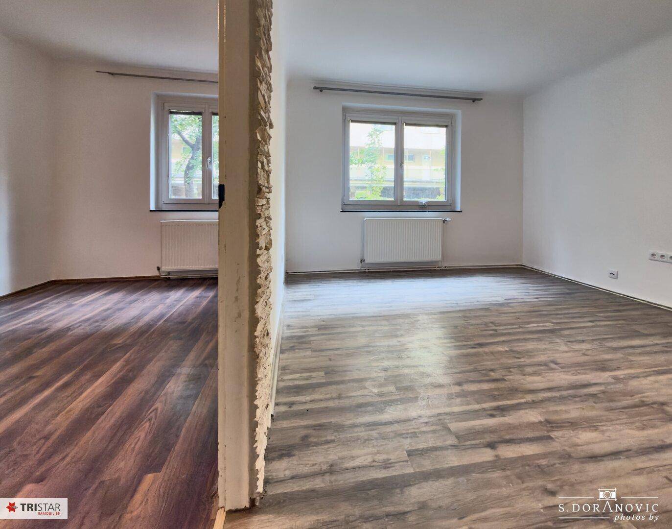 Helles Zimmer mit Laminatboden und zwei Fenstern, verbunden durch einen offenen Durchgang mit Steinakzenten.